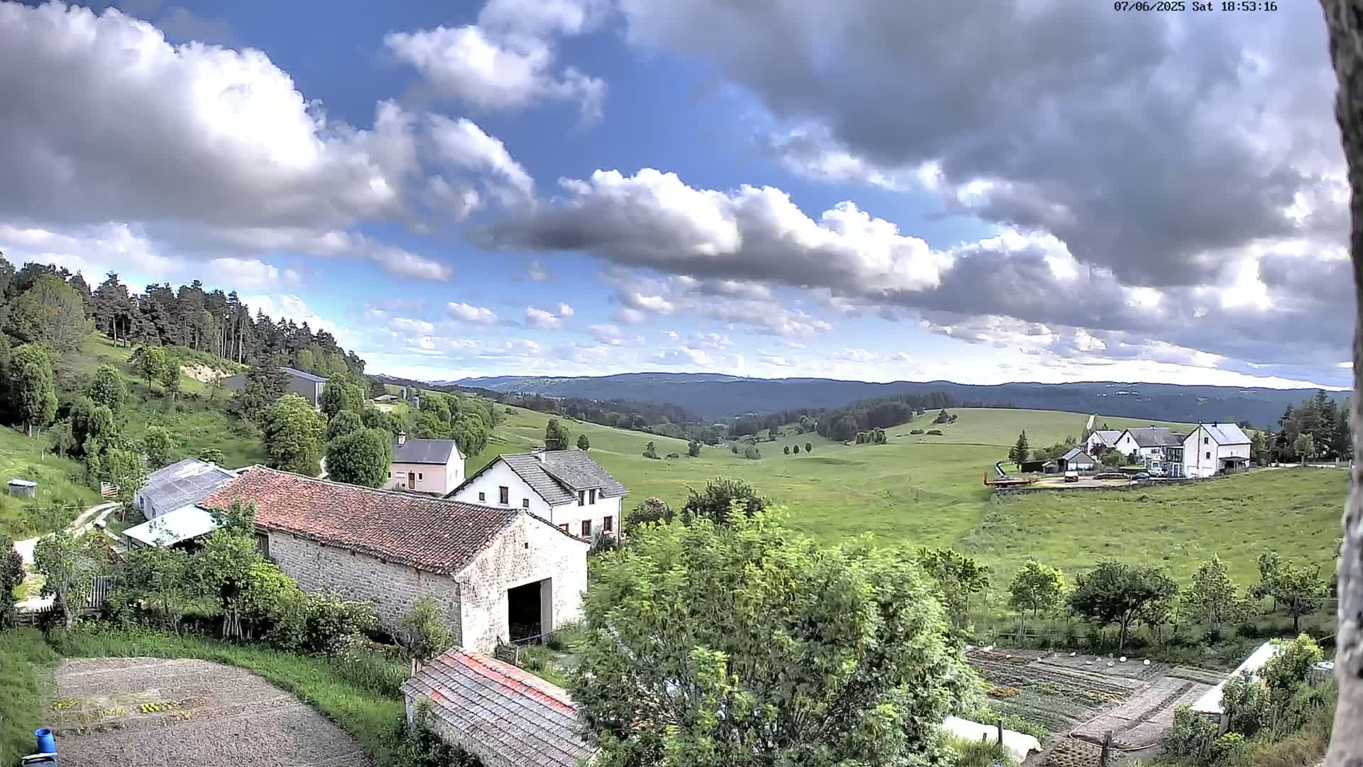 A partly cloudy day reveals a rural landscape with stone farmhouses, green pastures, and forested hills in the distance.