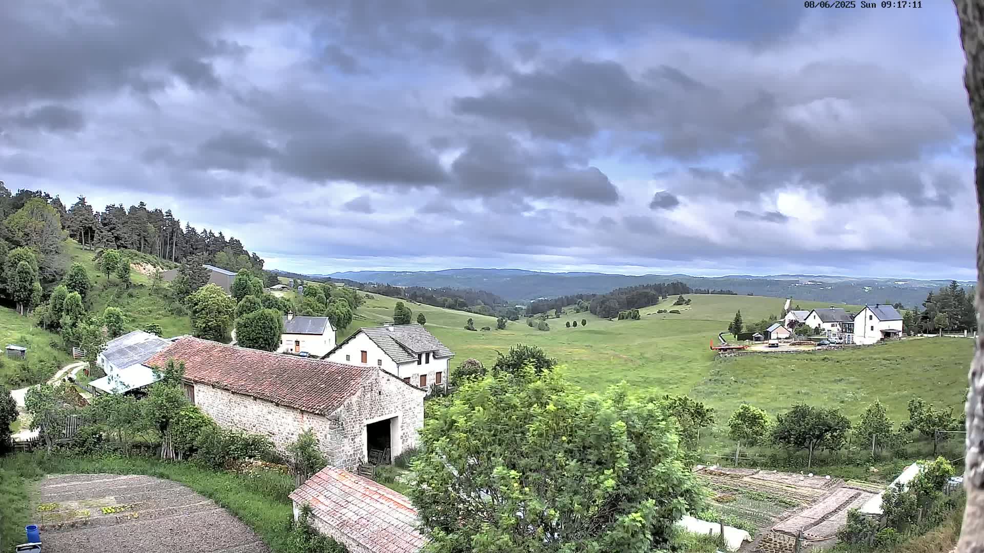A rural landscape under a cloudy sky shows stone buildings, green fields, and a forest in the distance.