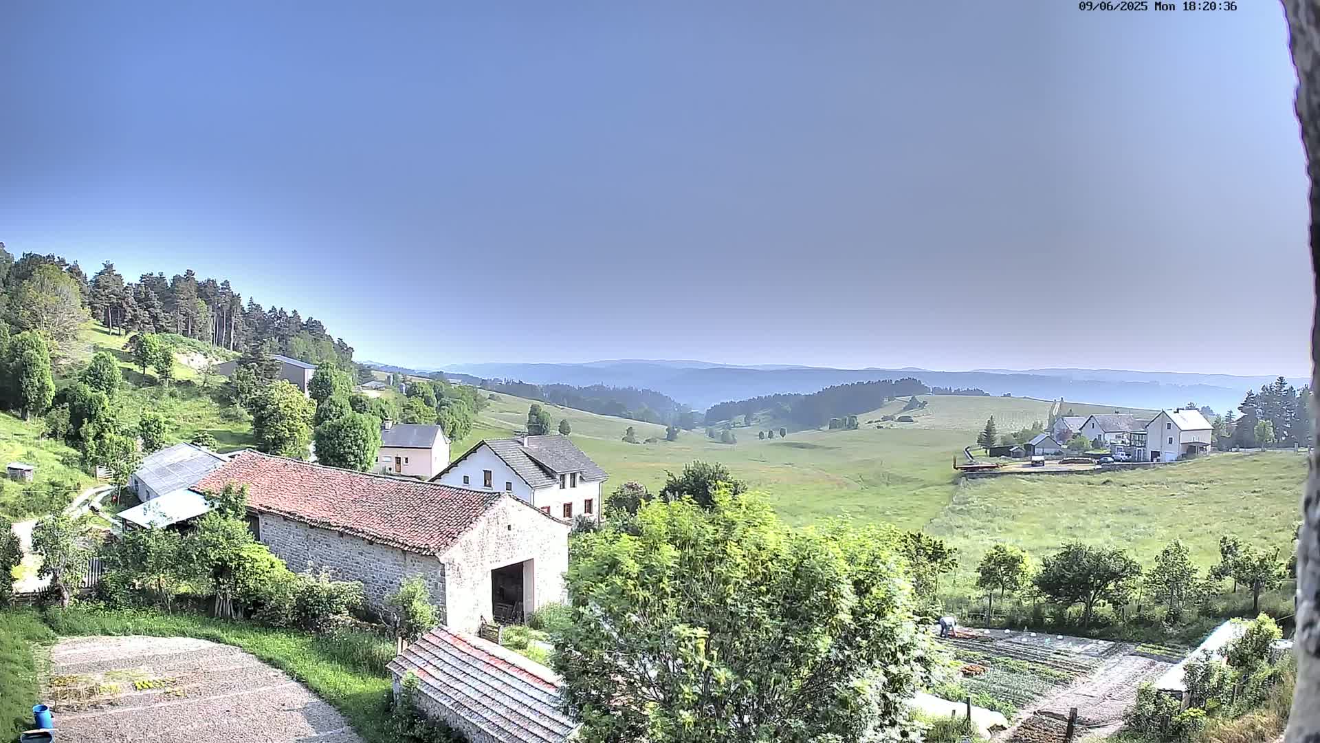 A high-angle view of a rural area under a clear blue sky shows several buildings nestled among green fields and trees, with distant rolling hills visible in the background.