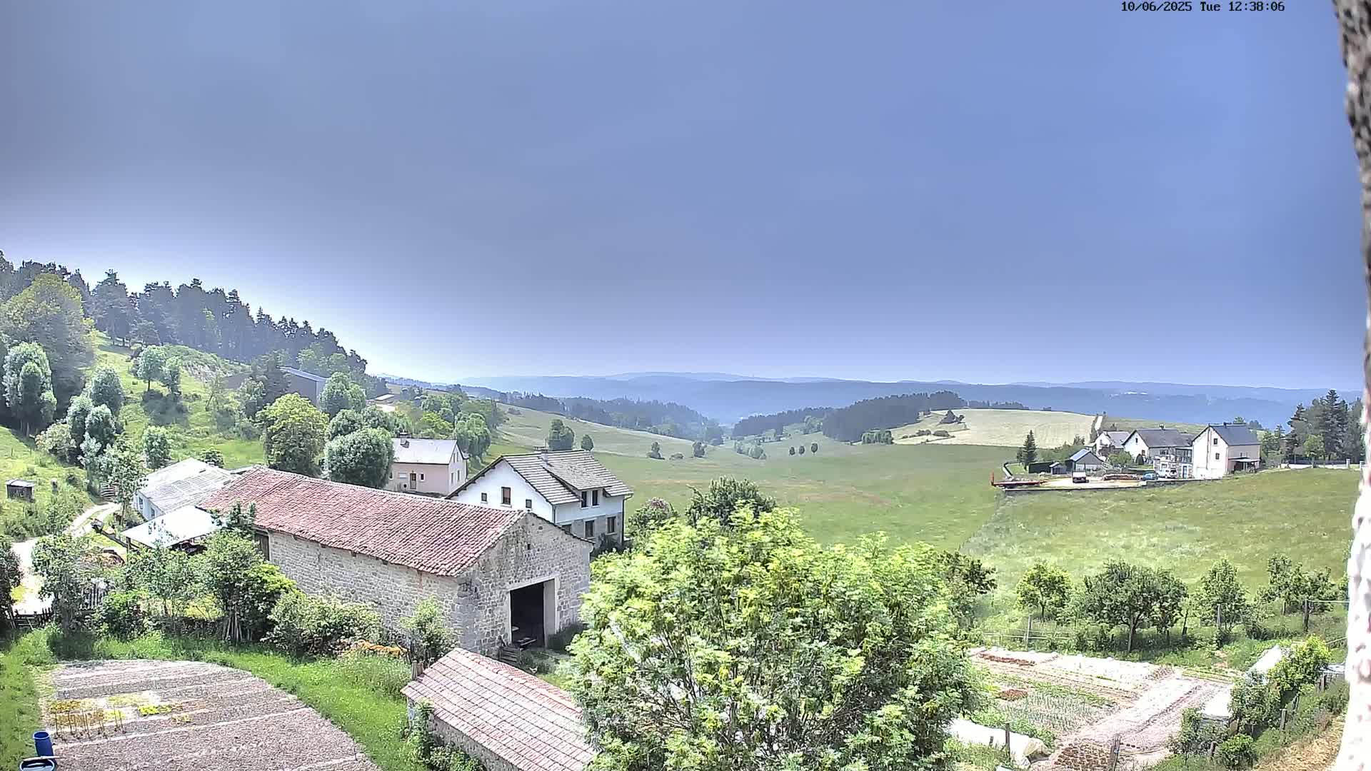 A rural village nestled in a valley, under a clear blue sky, is surrounded by green fields and forests.