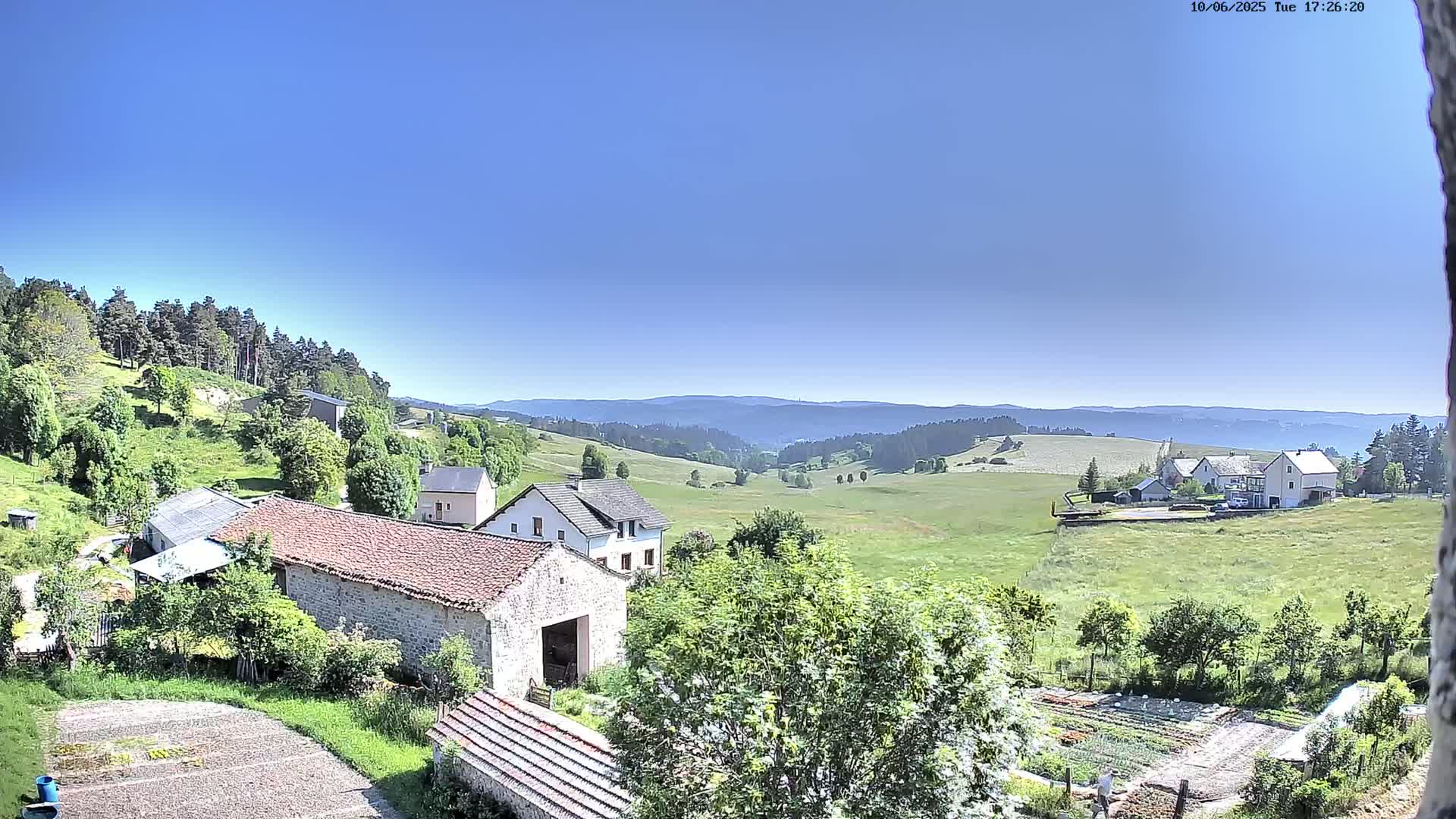 A rural landscape under a clear blue sky shows several buildings, green fields, and a forest-covered hill in the distance.