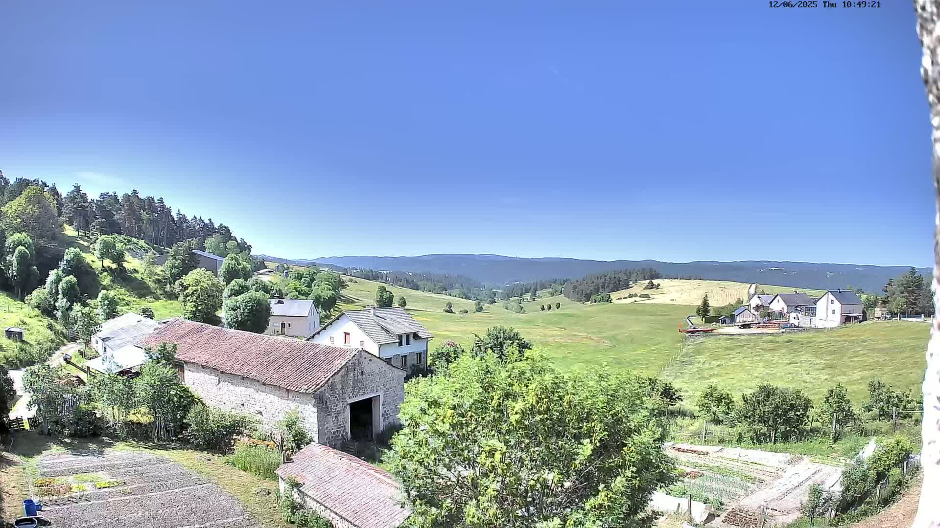 A sunny, clear day reveals a rural landscape of stone buildings, green fields, and distant forested hills.
