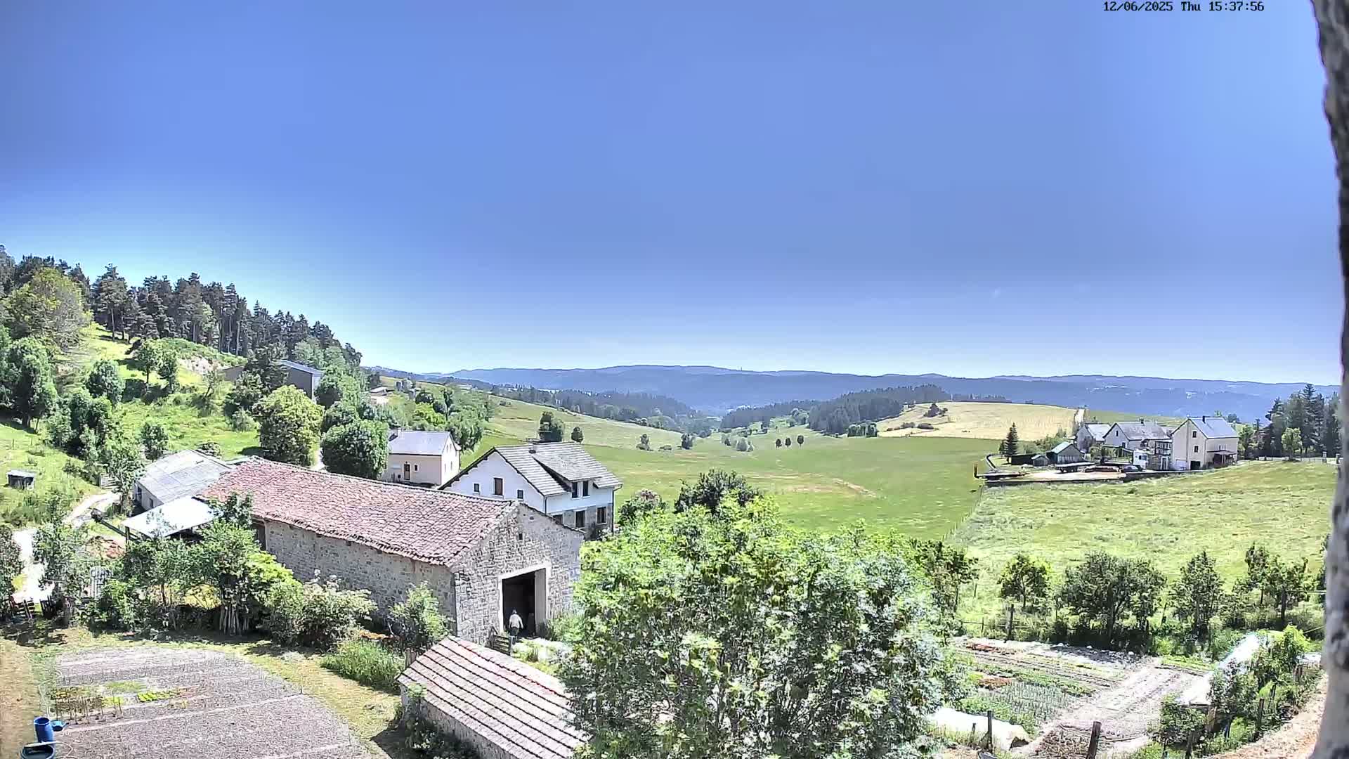 A sunny view of a rural village nestled in a valley, surrounded by green fields and forests under a clear blue sky.