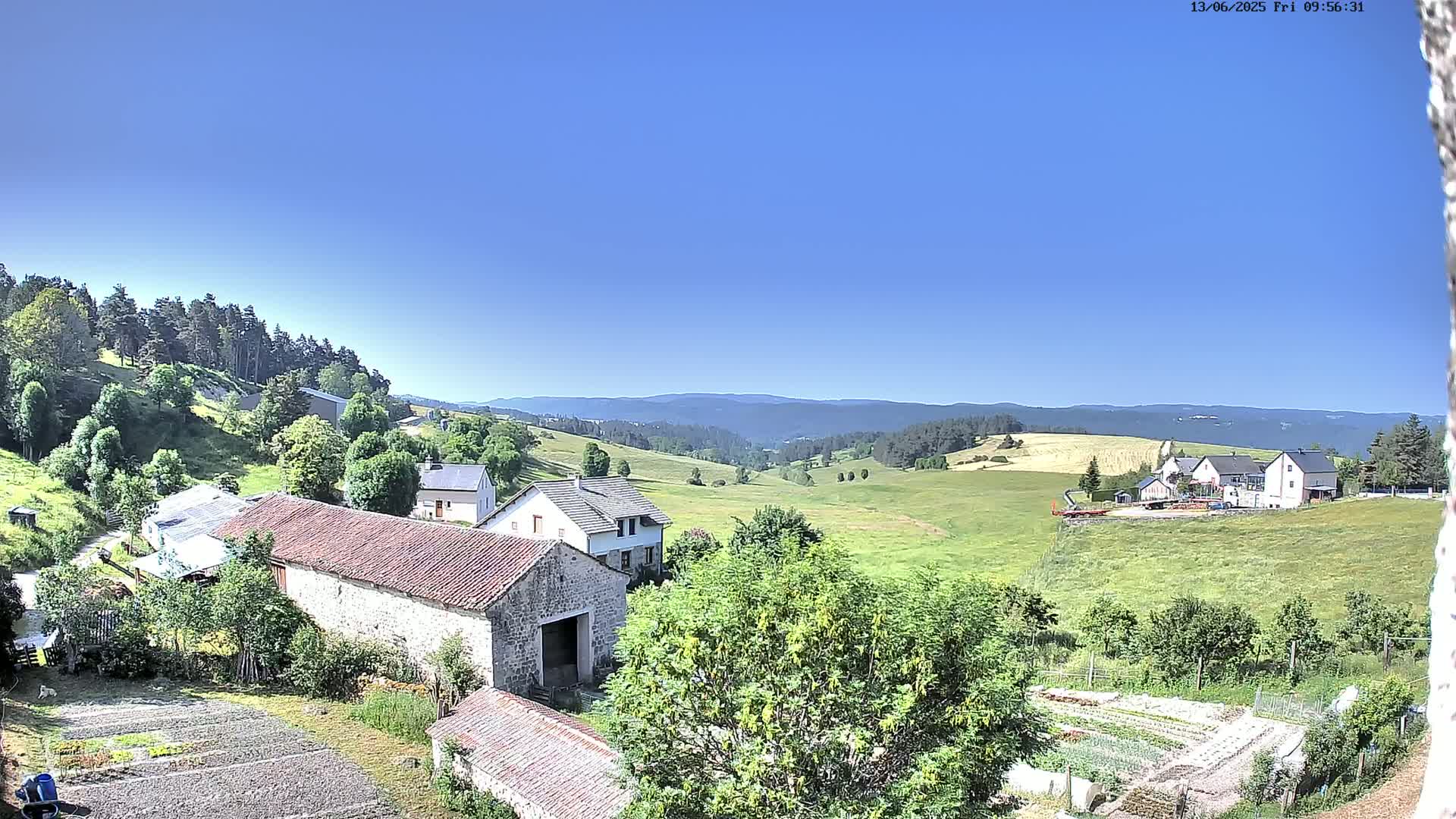 A rural scene on a clear, sunny day shows stone farm buildings, a green valley, and distant hills covered with forests.