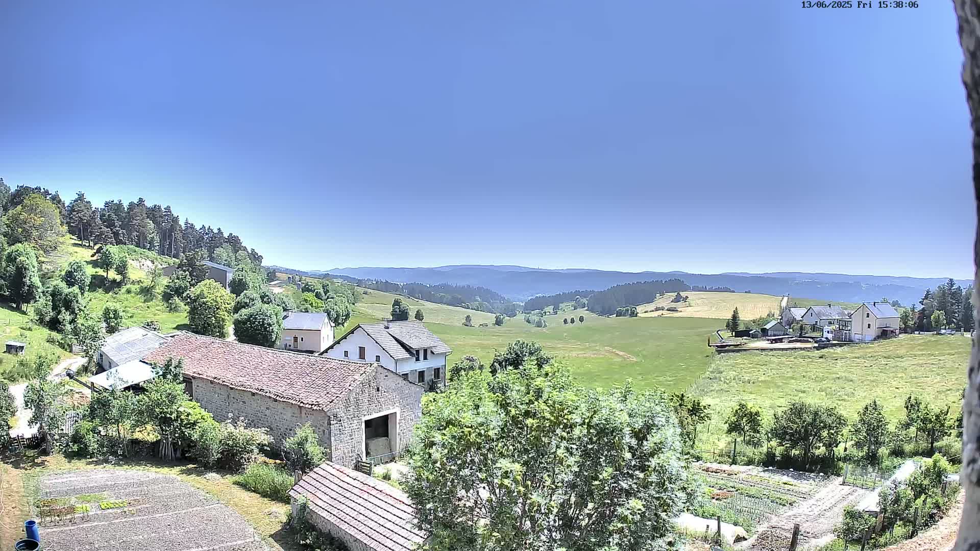 A sunny day reveals a rural landscape with stone buildings, green fields, and a distant mountain range under a clear blue sky.