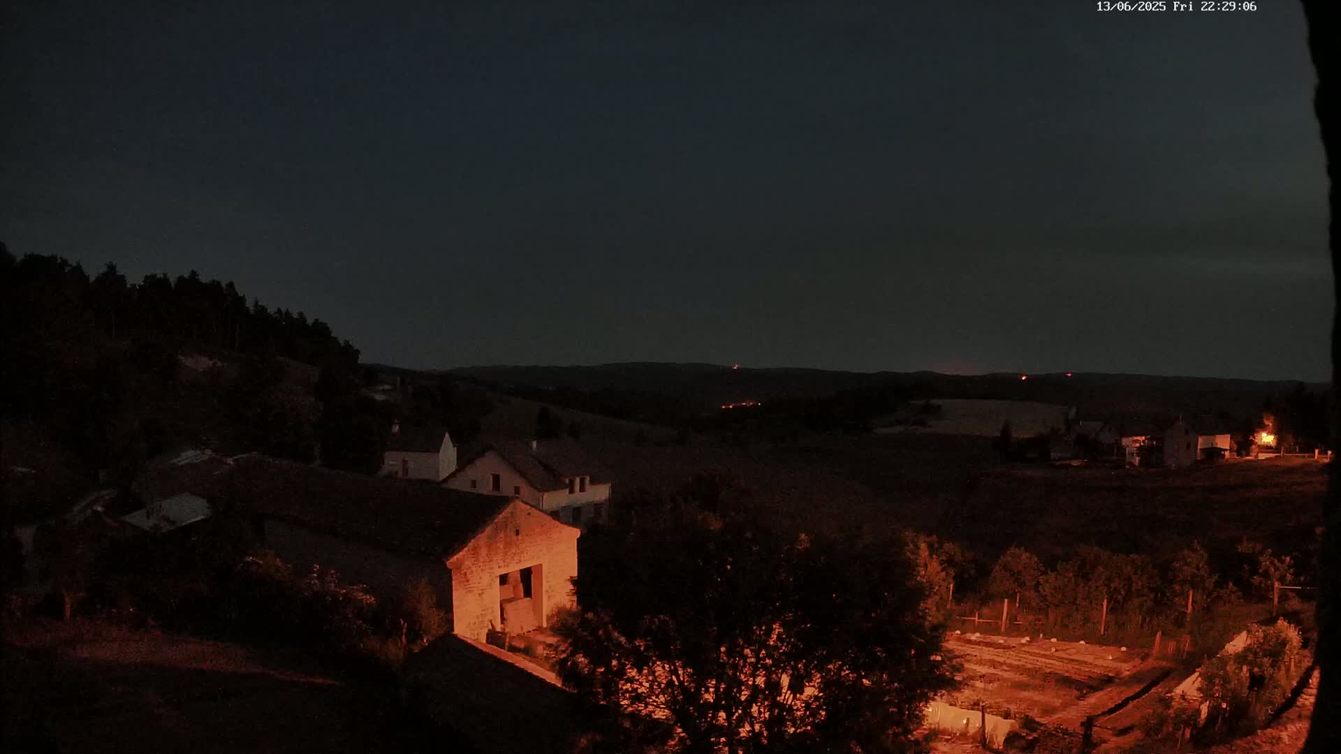 A nighttime view of a rural village nestled in a hilly landscape under a dark, clear sky shows several buildings illuminated with warm light.
