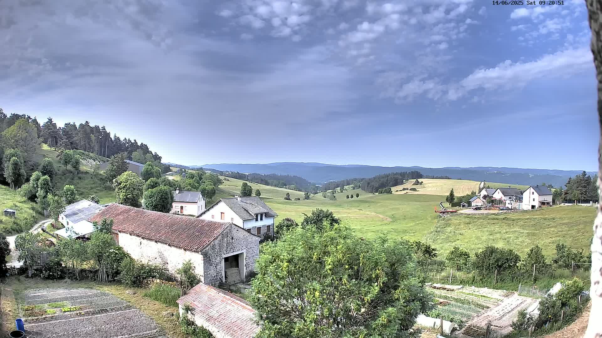 A partly cloudy sky overlooks a rural valley with several houses and farm buildings, green fields, and forested hills in the distance.