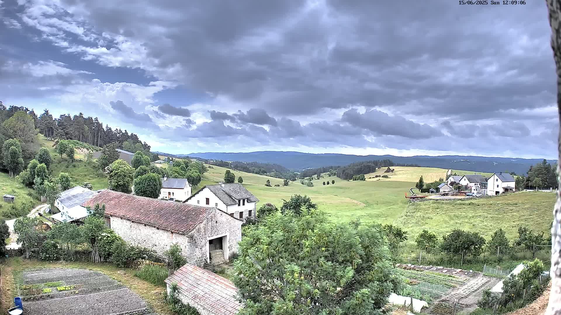 A rural landscape under a cloudy sky shows several buildings, farmlands, and forests extending into the distance.