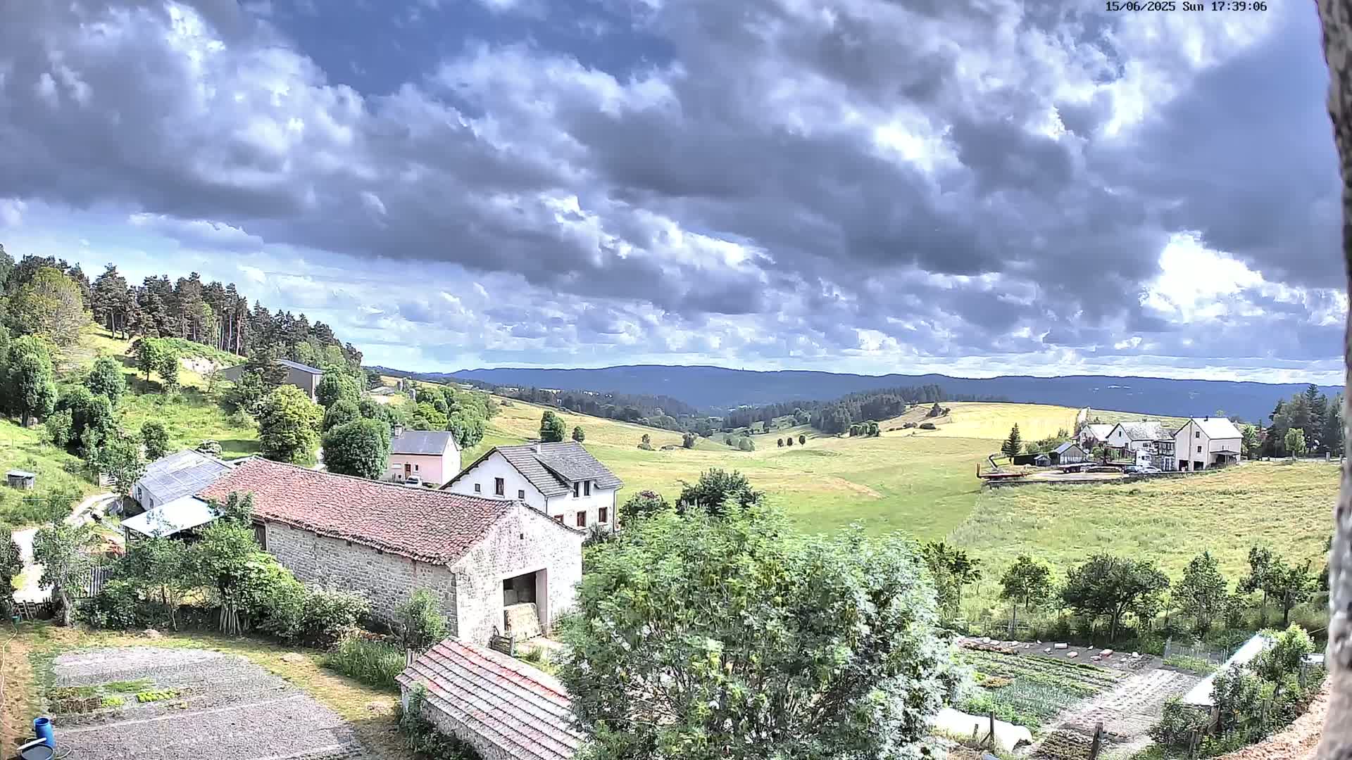 A rural landscape under a partly cloudy sky shows several stone buildings nestled among green fields and trees, with distant rolling hills in the background.