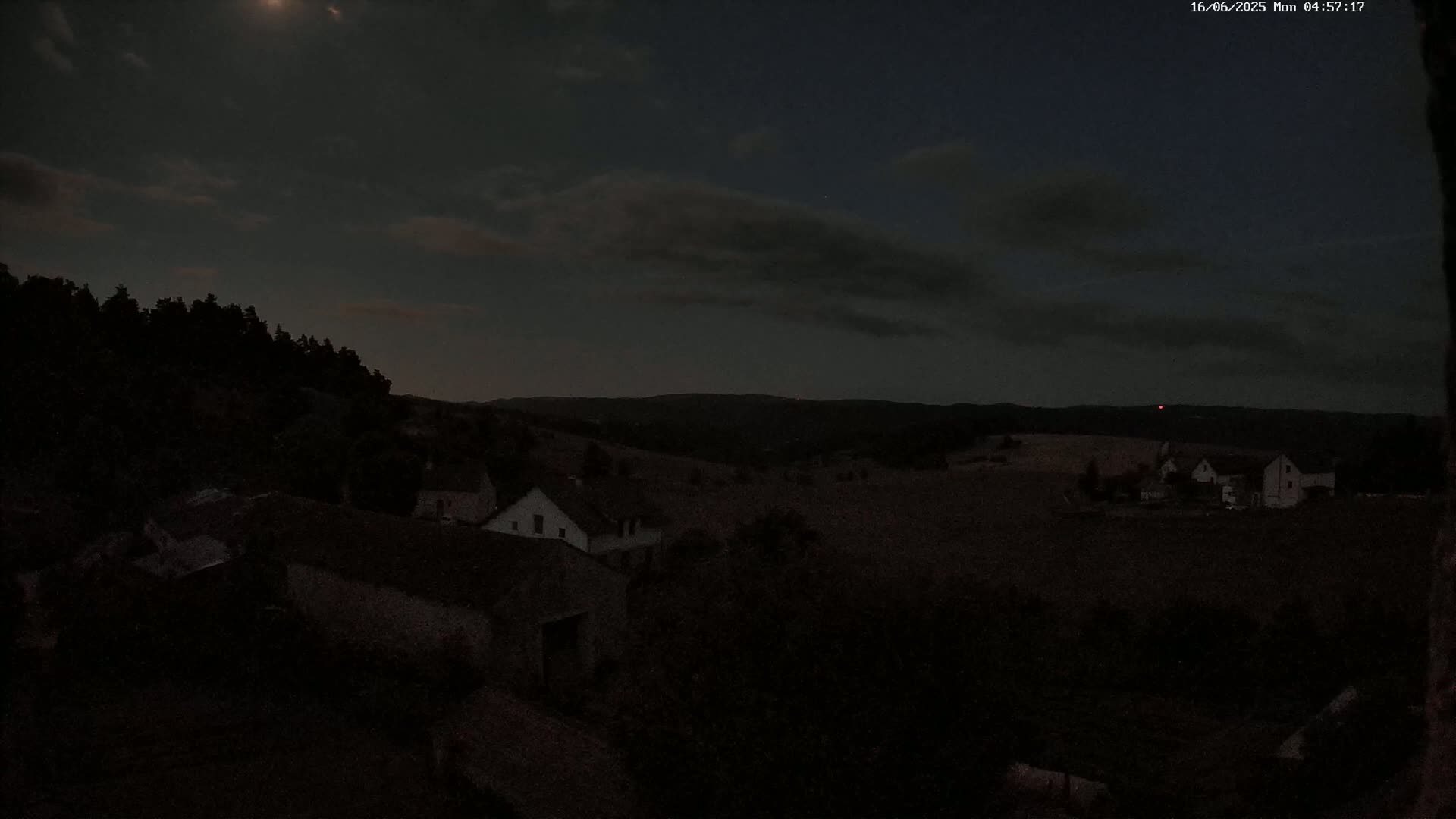 A moonlit, nighttime scene shows a few buildings nestled in a dark, rural landscape under a partly cloudy sky.