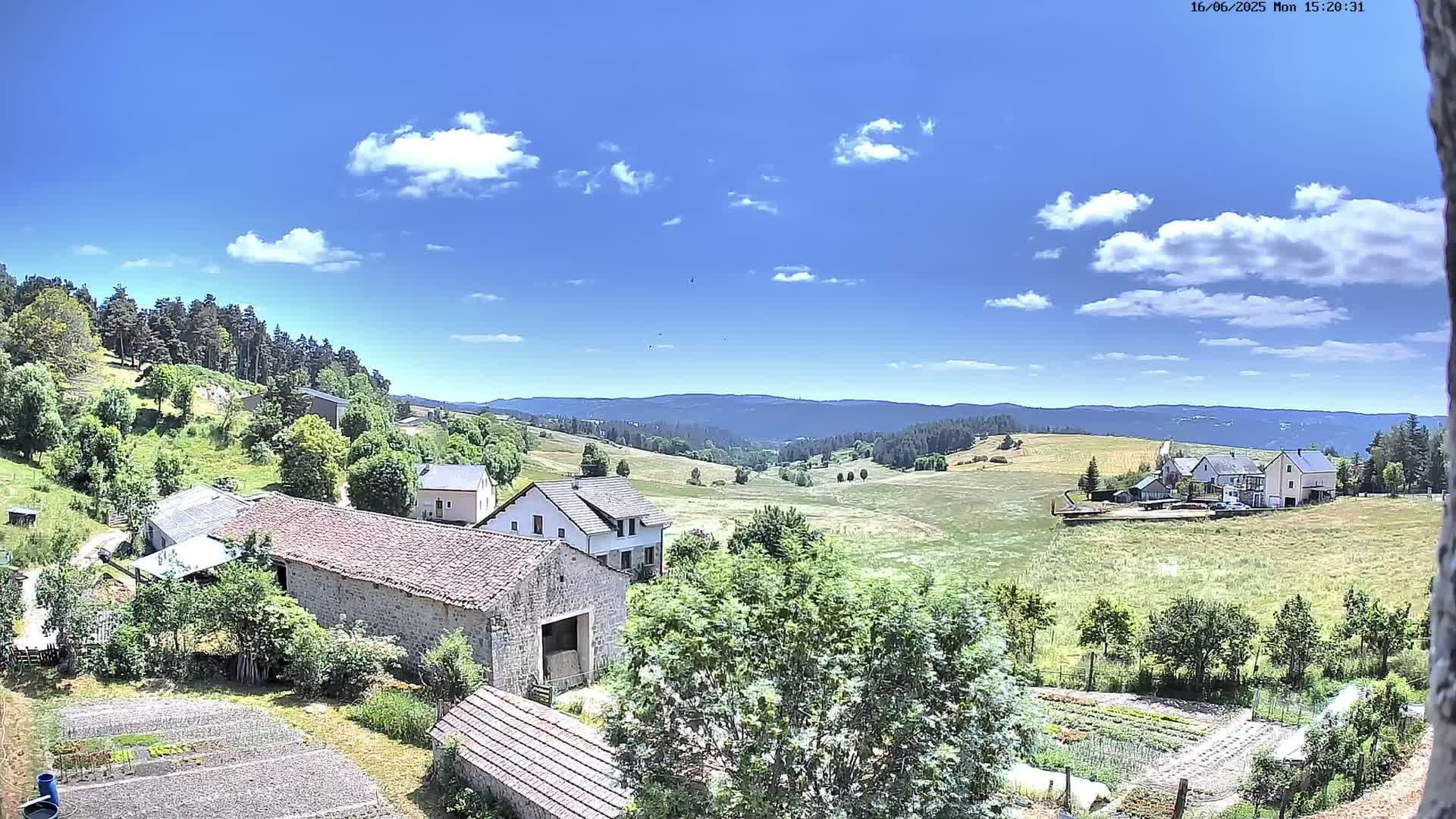 A sunny day with scattered clouds reveals a rural landscape of houses, farm buildings, and fields, nestled amidst rolling hills and forests.