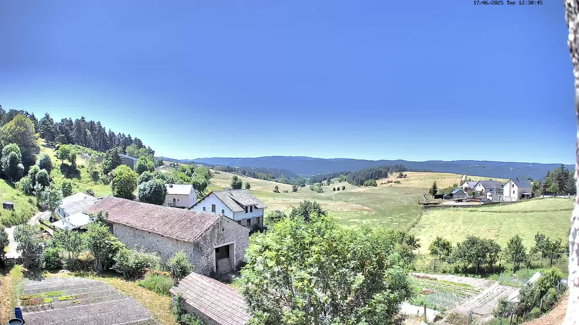 A sunny day reveals a rural landscape of stone buildings, green fields, and forested hills under a clear blue sky.