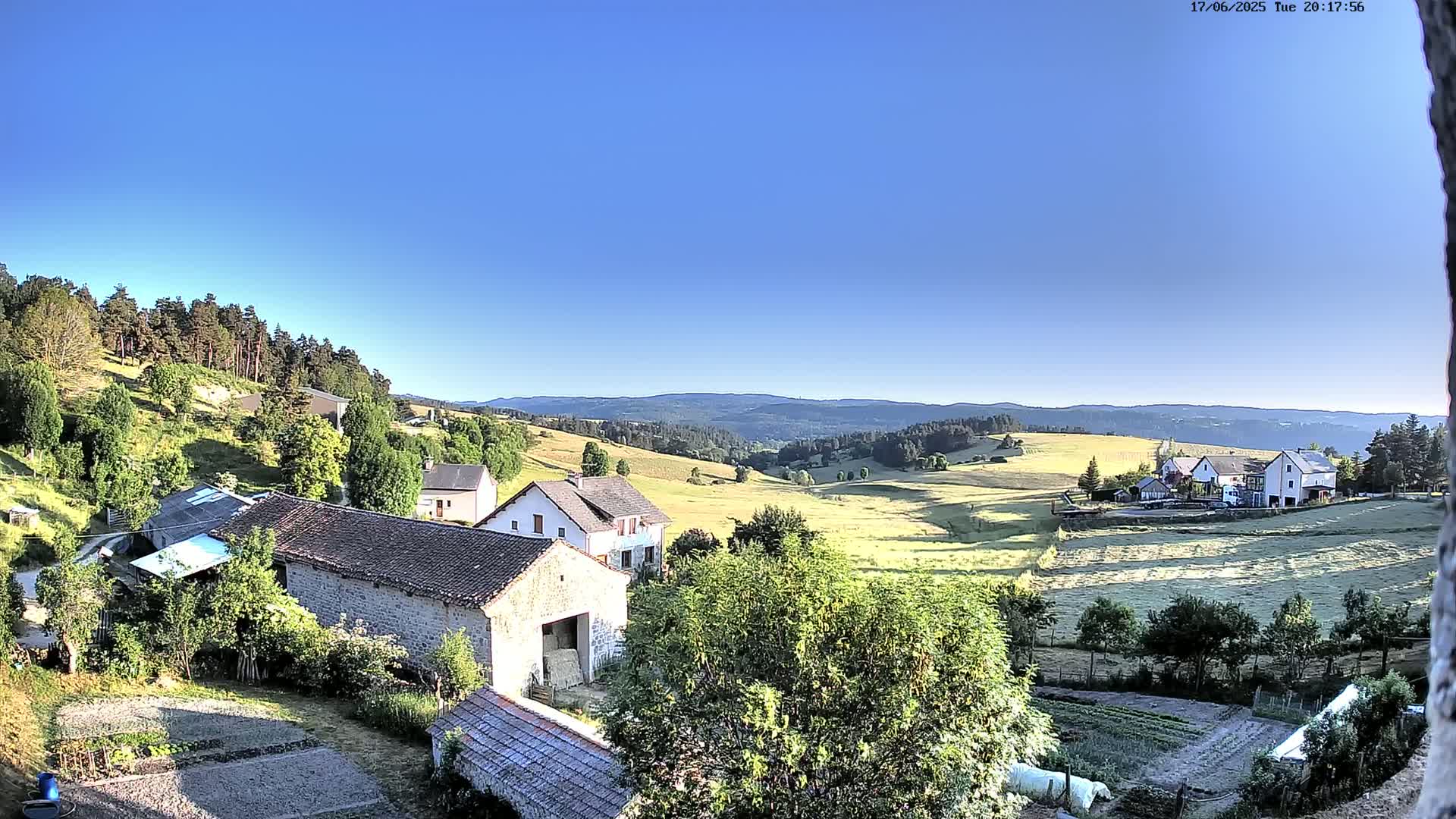 A high-angle view shows a rural landscape of stone buildings, fields, and forests under a clear blue sky.