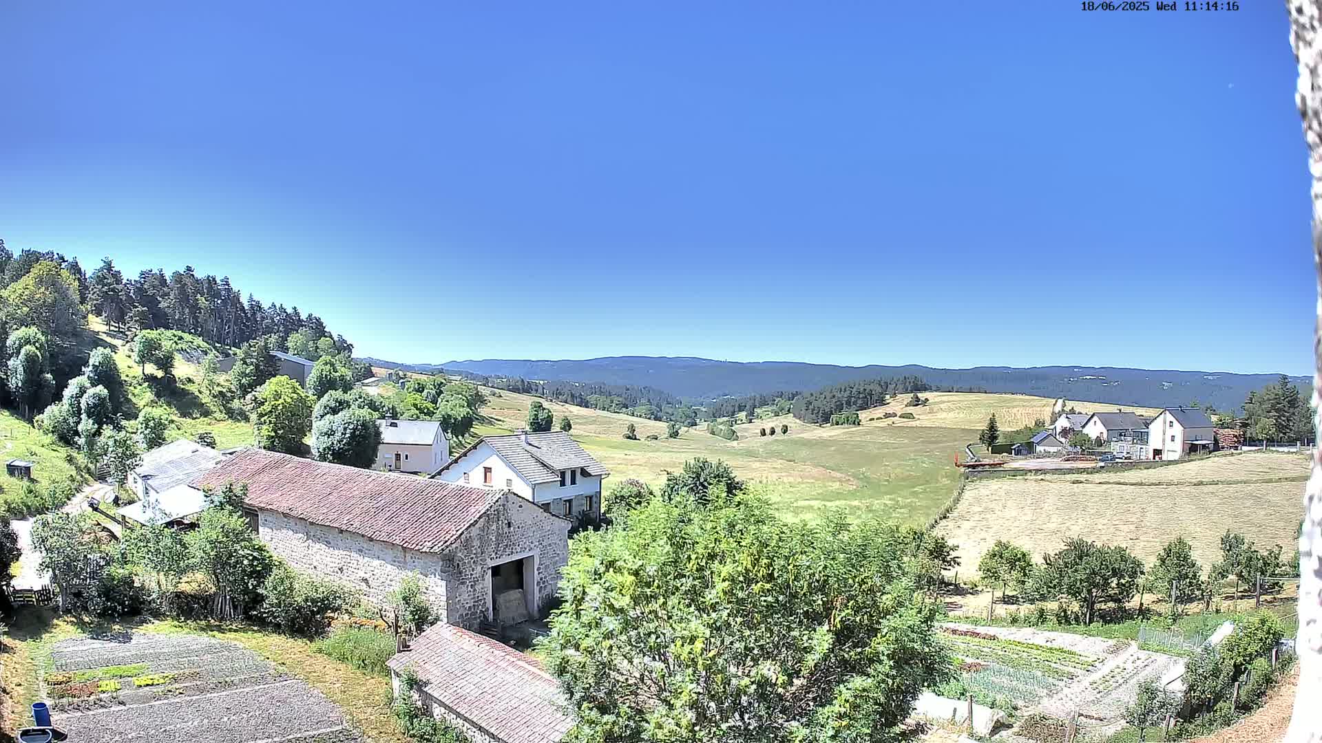 A sunny, clear-day view of a rural valley with stone buildings, farmlands, and a forest in the distance.