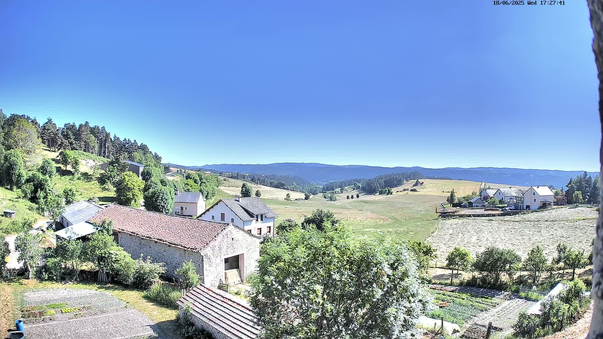 A high-angle view of a rural landscape under a clear blue sky shows several stone buildings, green fields, and a distant mountain range.