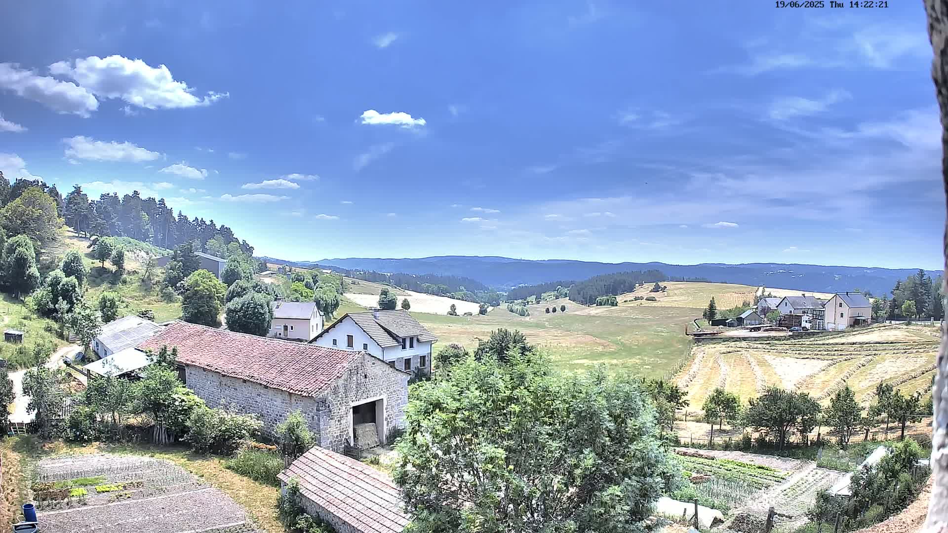 A sunny day reveals a rural landscape with stone farmhouses, cultivated fields, and a background of rolling hills under a partly cloudy sky.