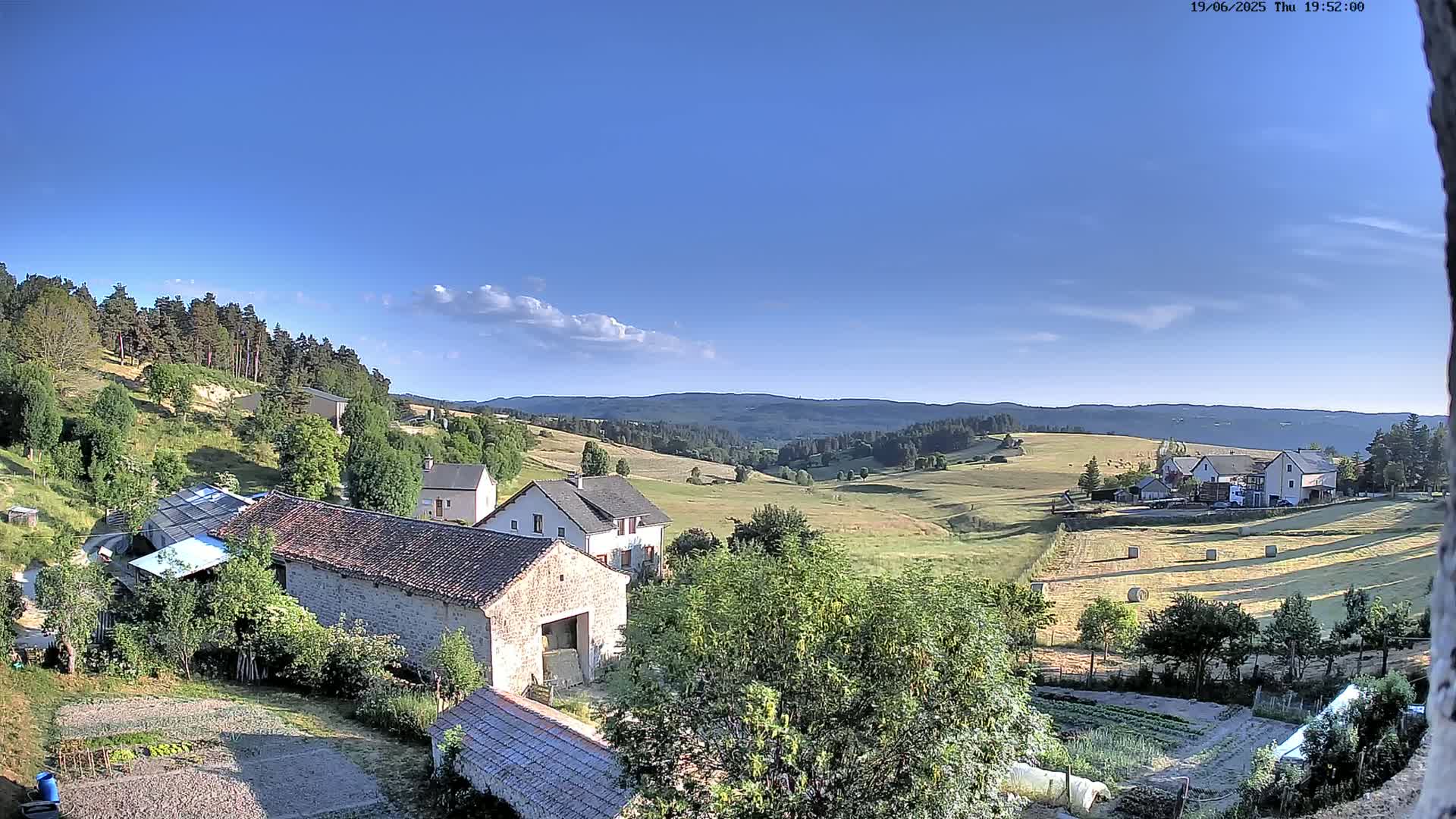 A high-angle view of a rural landscape under a mostly clear blue sky shows several stone buildings and farm structures nestled amongst green hills and fields, with hay bales visible in the distance.