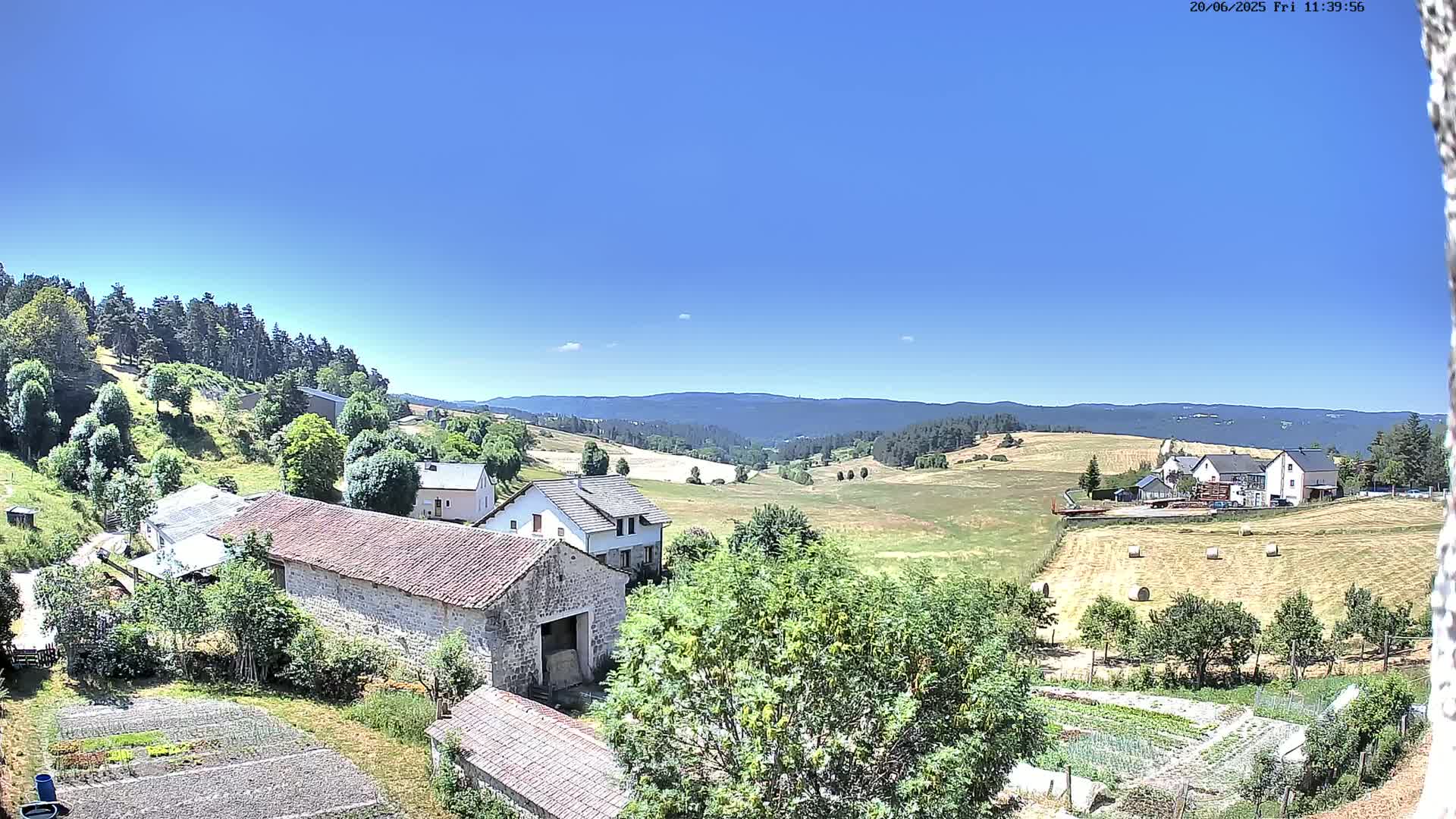 Stone farm buildings and cultivated gardens sit nestled in a verdant valley under a clear blue sky, with distant hills covered in forests.