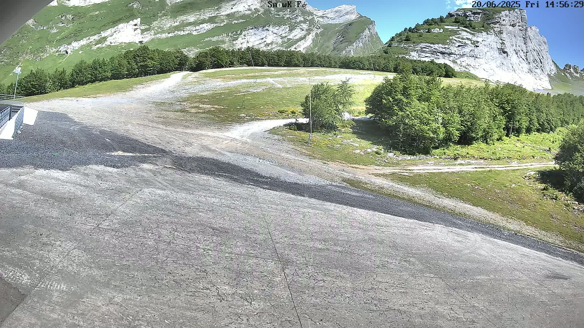 A gray, sloped paved area leads to a grassy field and a rocky mountainside under a mostly sunny sky.