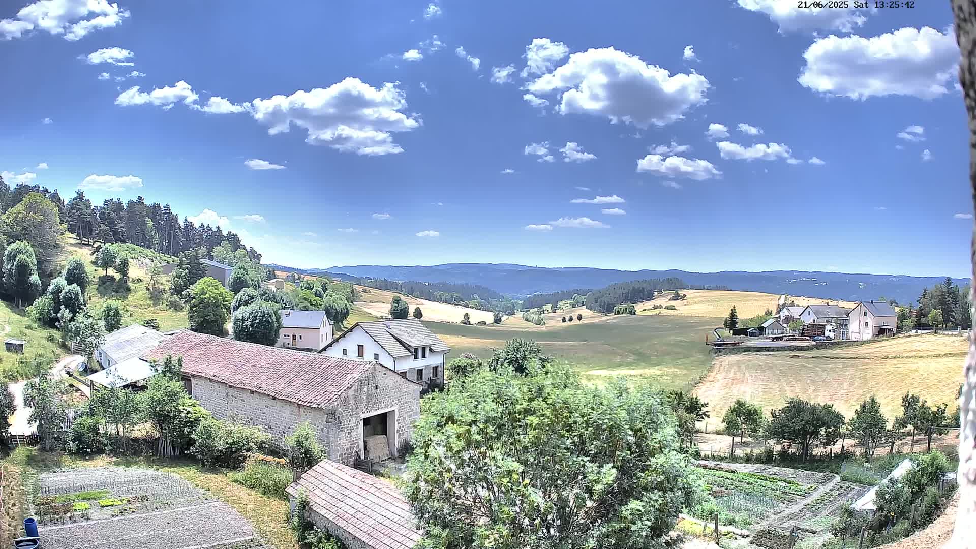 A sunny day reveals a rural landscape with stone farmhouses, cultivated fields, and a background of rolling hills covered in forests under a partly cloudy sky.