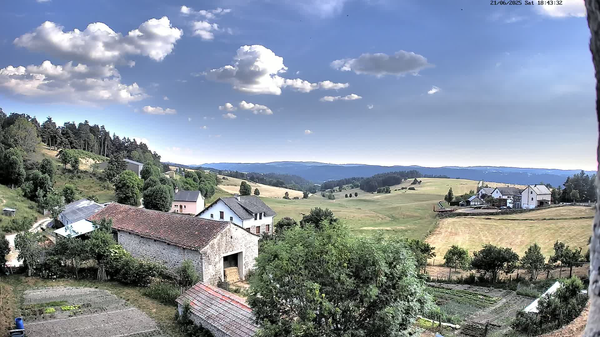 A partly cloudy sky overlooks a rural landscape of houses, farm buildings, and fields, with a distant line of forested hills.
