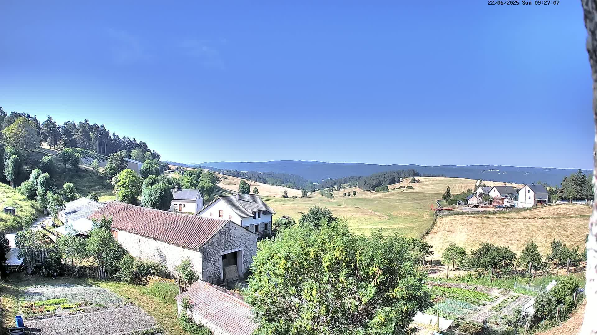 A sun-drenched rural landscape displays stone farm buildings and gardens nestled among green hills and distant mountains under a clear blue sky.