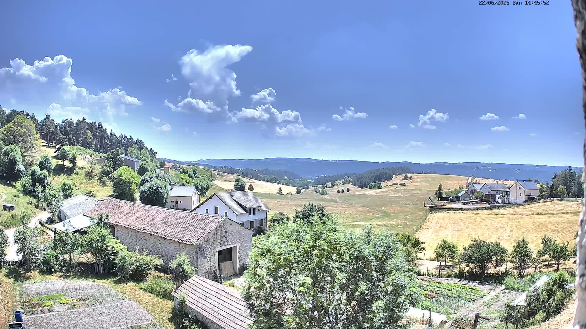 A sunny day reveals a rural landscape with stone buildings, farm fields, and a background of rolling hills covered in forest.