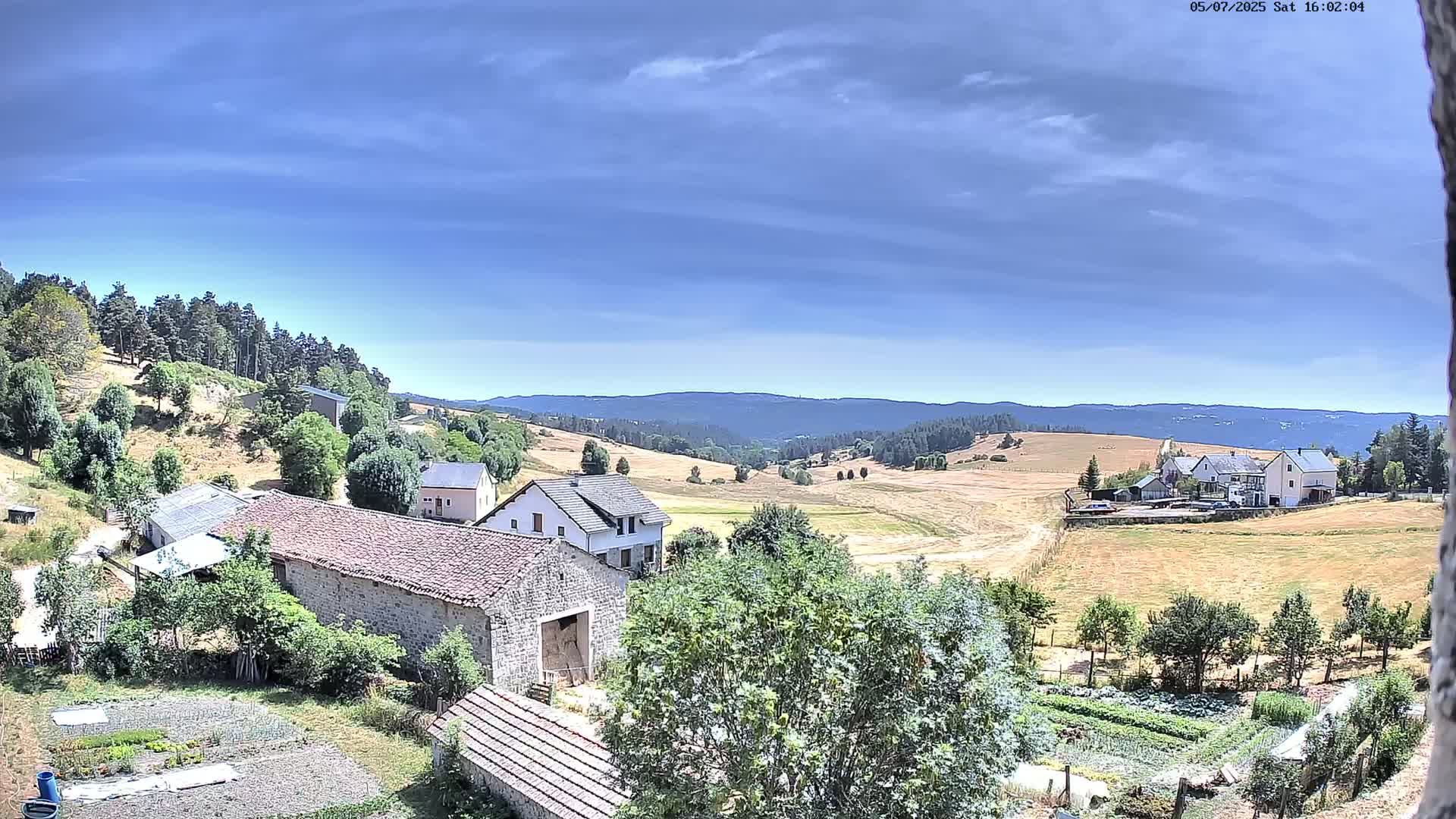 A partly cloudy sky overlooks a rural landscape of stone buildings, farmlands, and a forest-covered hill under sunny conditions.
