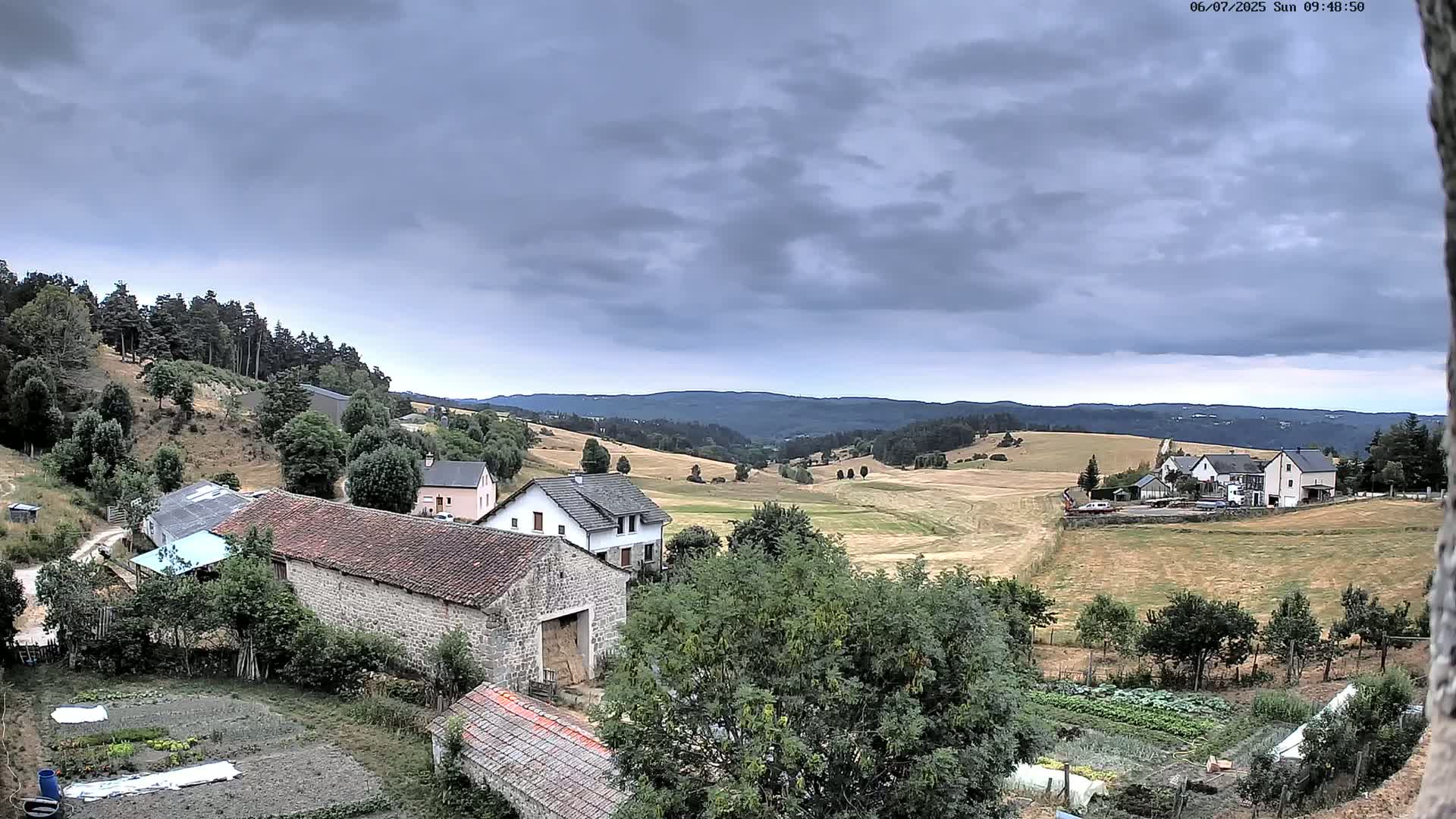 A rural landscape under a cloudy sky shows several stone buildings, farmlands, and gardens nestled among rolling hills and distant forests.