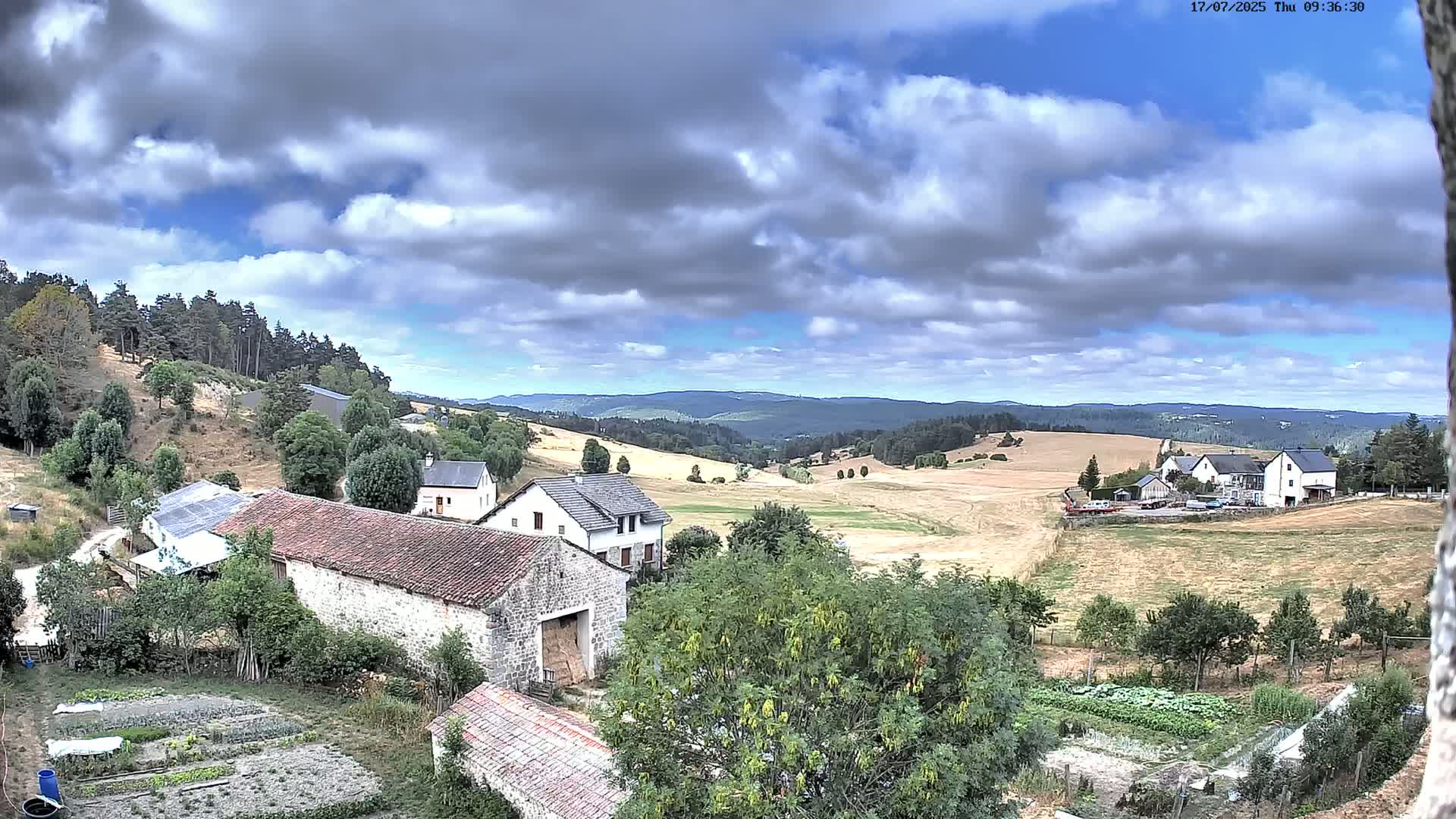 A partly cloudy day reveals a rural landscape with stone buildings, farm fields, and a distant line of hills.
