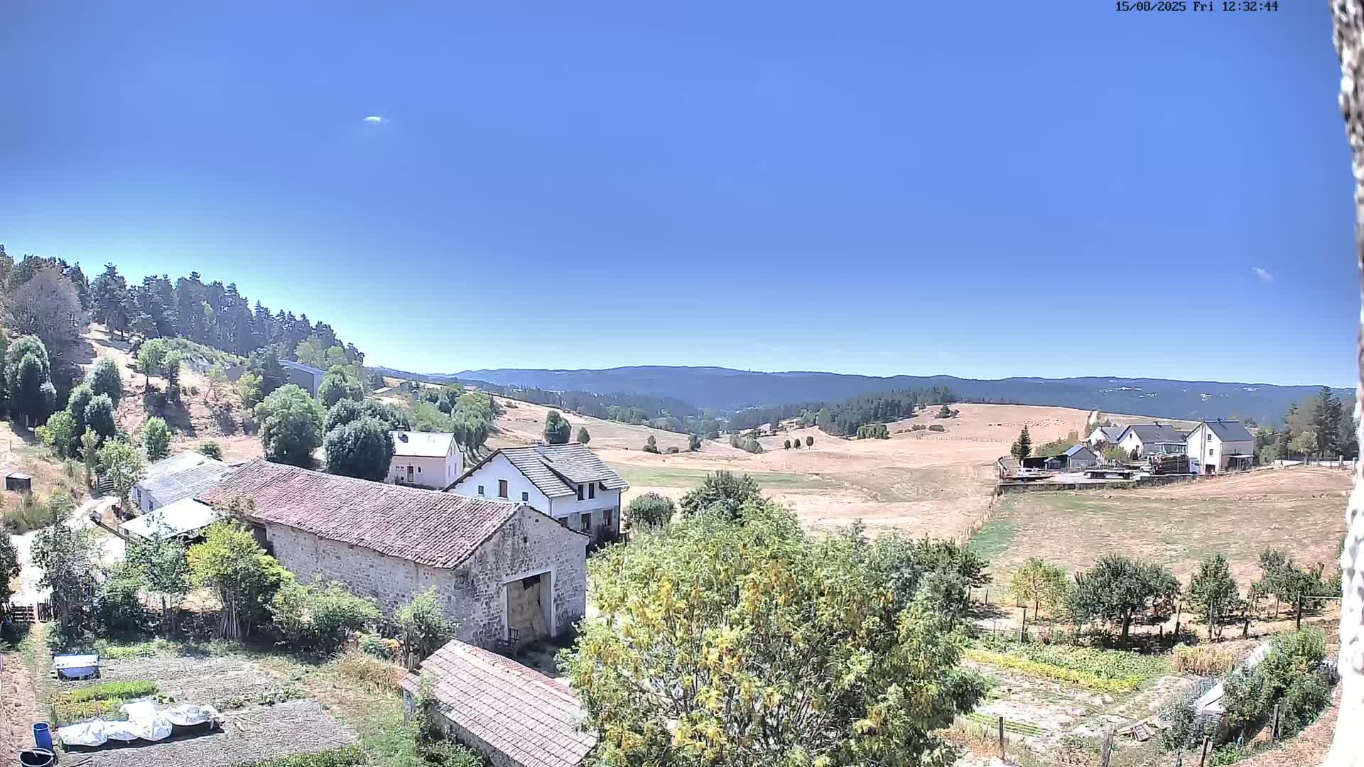 A partly cloudy day reveals a rural landscape with stone buildings, farm fields, and a distant line of hills.