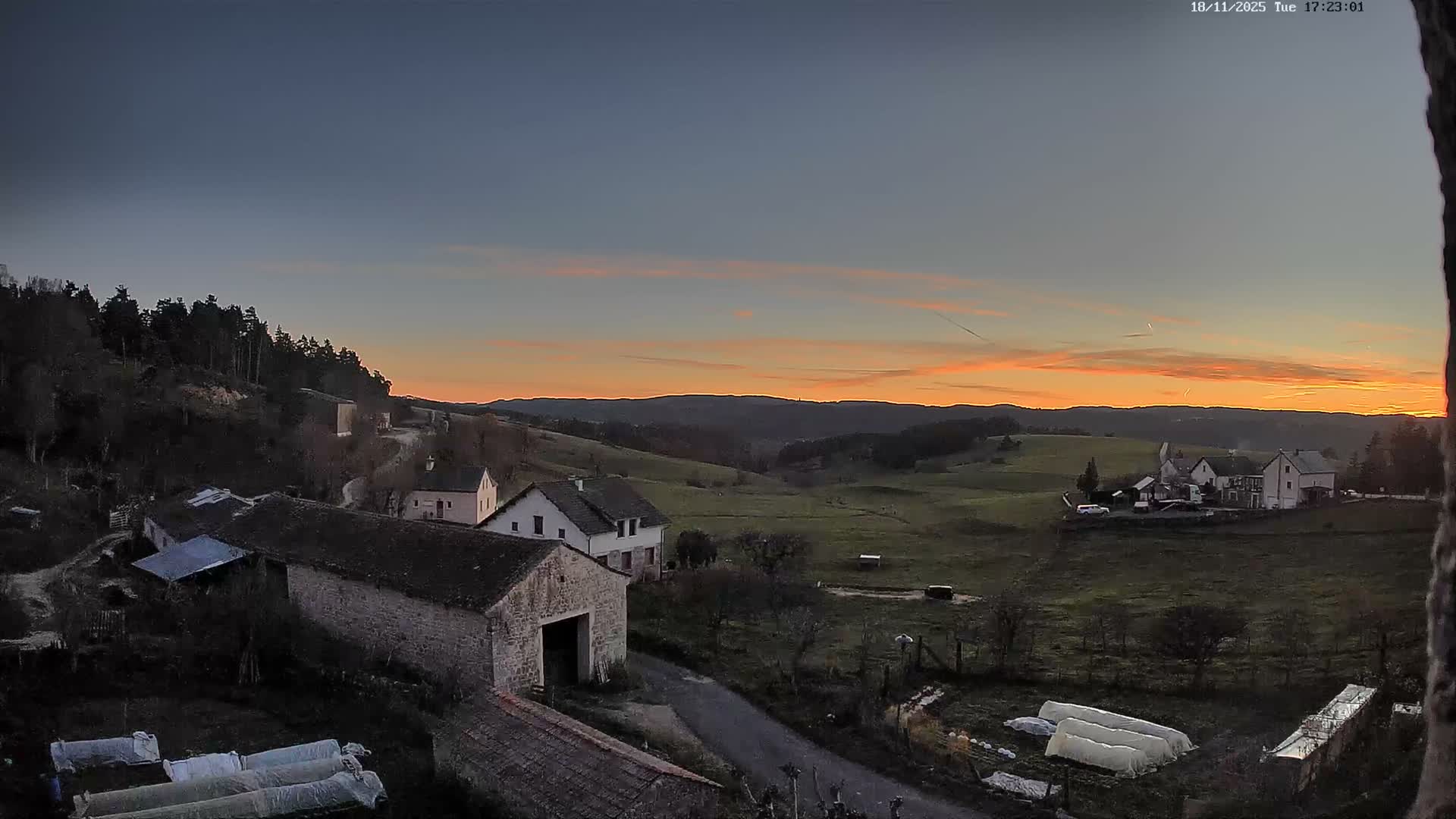 A partly cloudy day reveals a rural landscape with stone buildings, farm fields, and a distant line of hills.