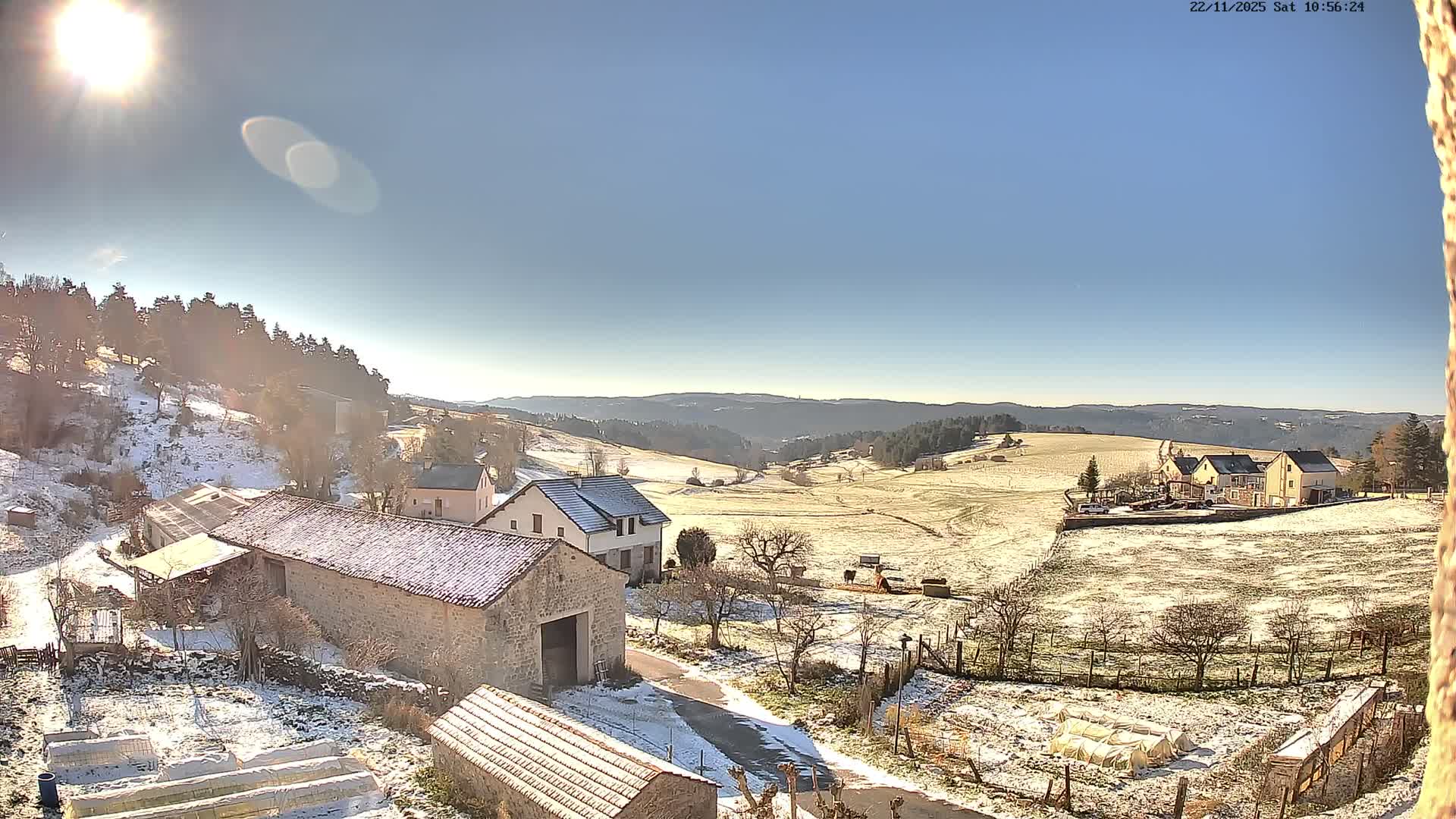 A bright, sunny winter day illuminates a serene rural landscape where light snow covers stone houses, farm buildings, fields, and bare trees, stretching towards distant rolling hills under a clear blue sky.