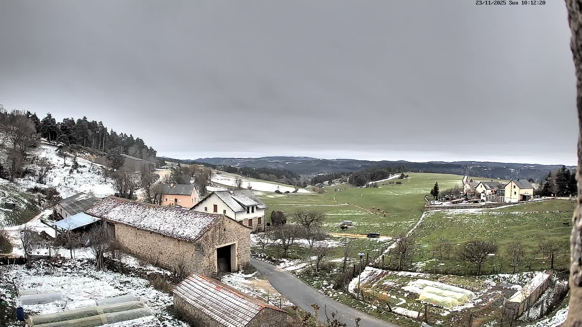 The image depicts a rural landscape lightly dusted with snow on the ground and rooftops, revealing green fields and scattered buildings under a uniformly grey, overcast sky.
