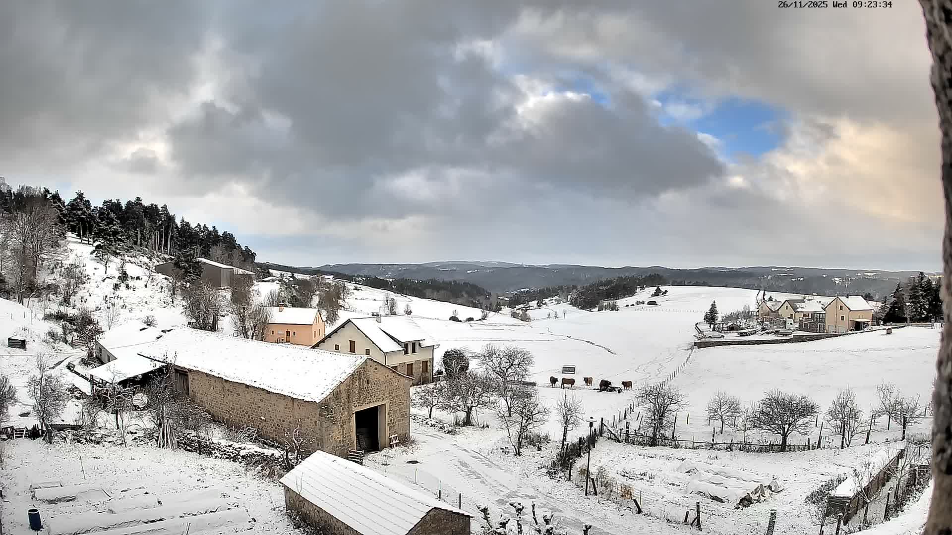A snow-covered rural village and surrounding rolling hills are seen under a partly cloudy sky with hints of blue and sunlight, featuring scattered houses, bare and evergreen trees, and a few cows in a fenced field.