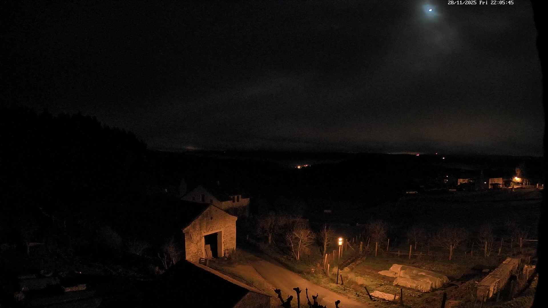 A dark, partly cloudy night illuminates a rural valley featuring stone houses, a winding road with a streetlight, and distant lights under a visible crescent moon.