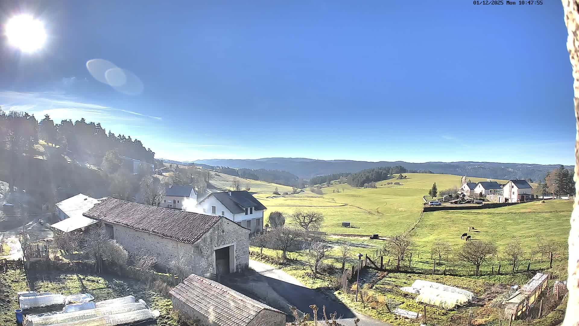 A bright, clear, and sunny day illuminates a frosty rural landscape featuring traditional stone buildings, white houses, green pastures with grazing cows, and distant forested hills under a brilliant blue sky.