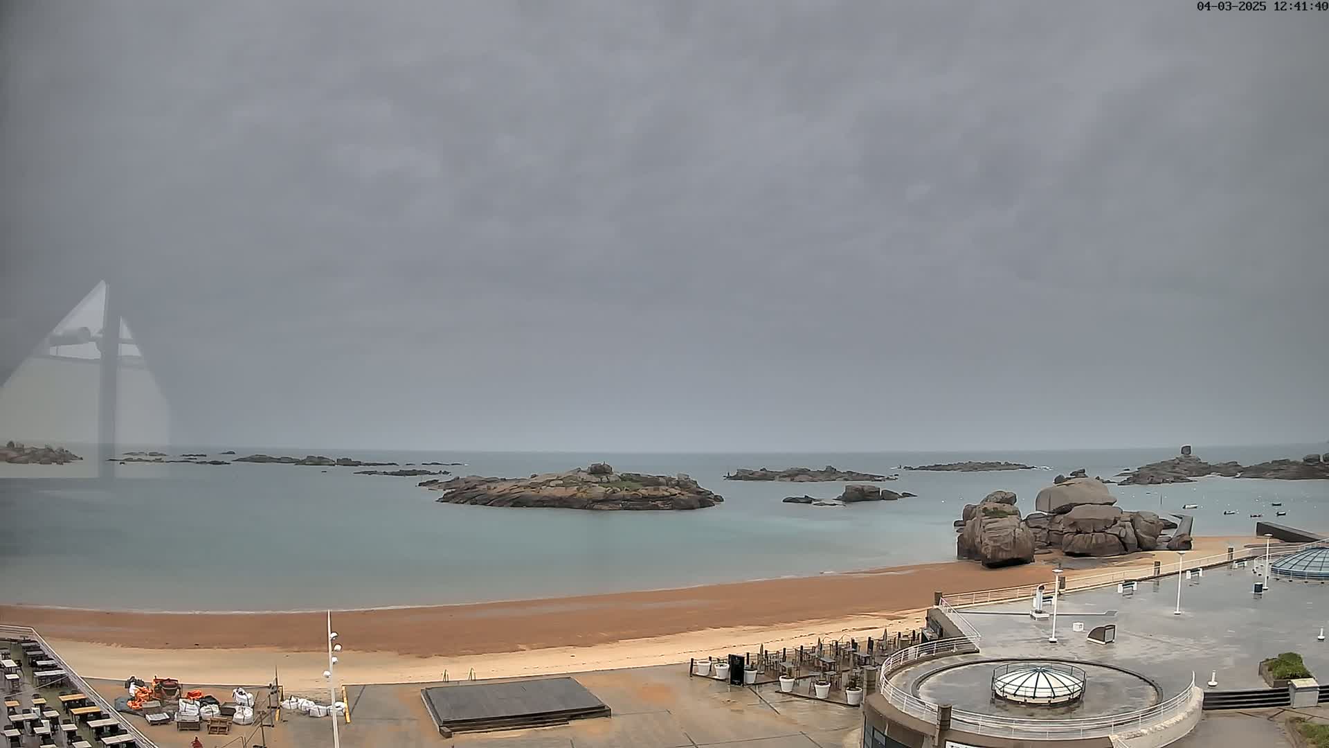 A sandy beach with rocky islands in a calm, overcast sea is viewed from above a seaside building.