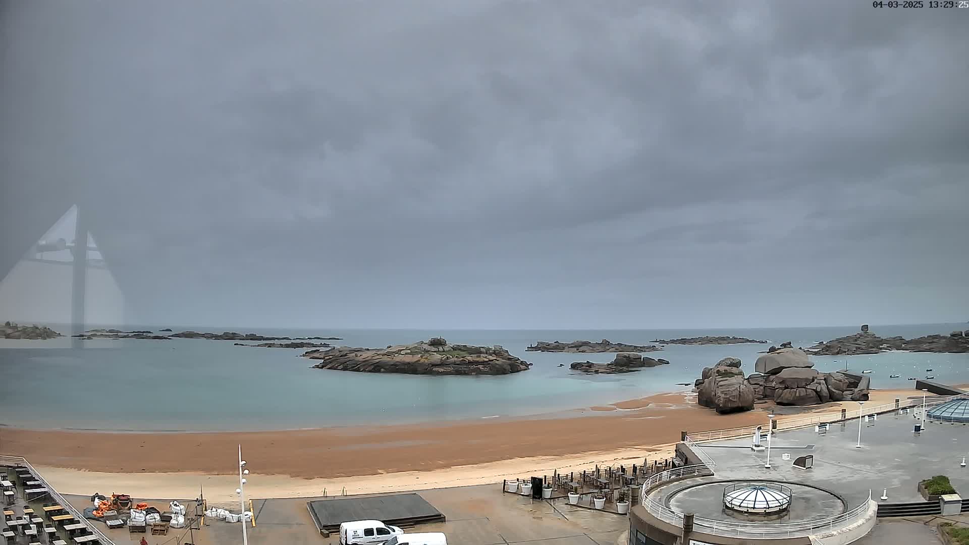 A sandy beach with rocky outcrops in a calm, overcast body of water is viewed from above under a gray, cloudy sky.