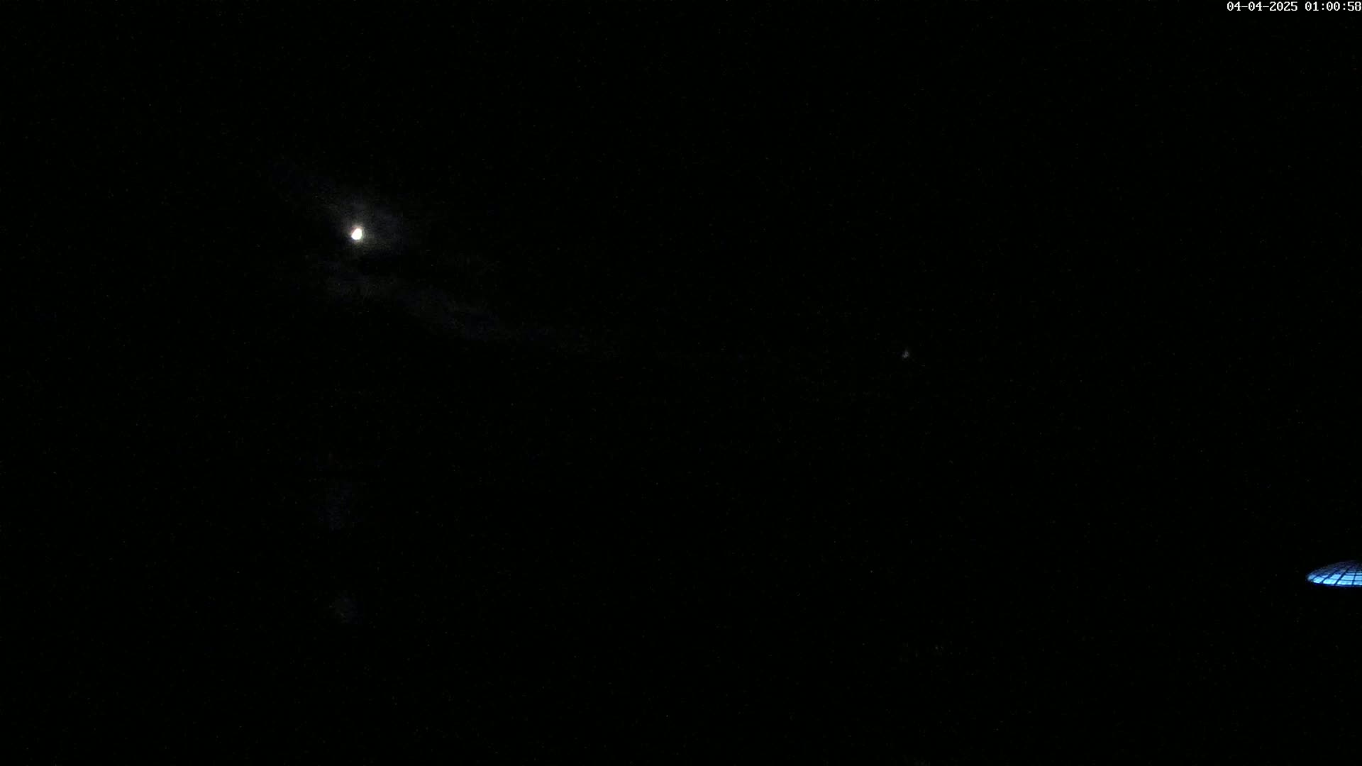 A nighttime outdoor scene shows a bright moon partially obscured by clouds and a small portion of a satellite in the lower right corner.