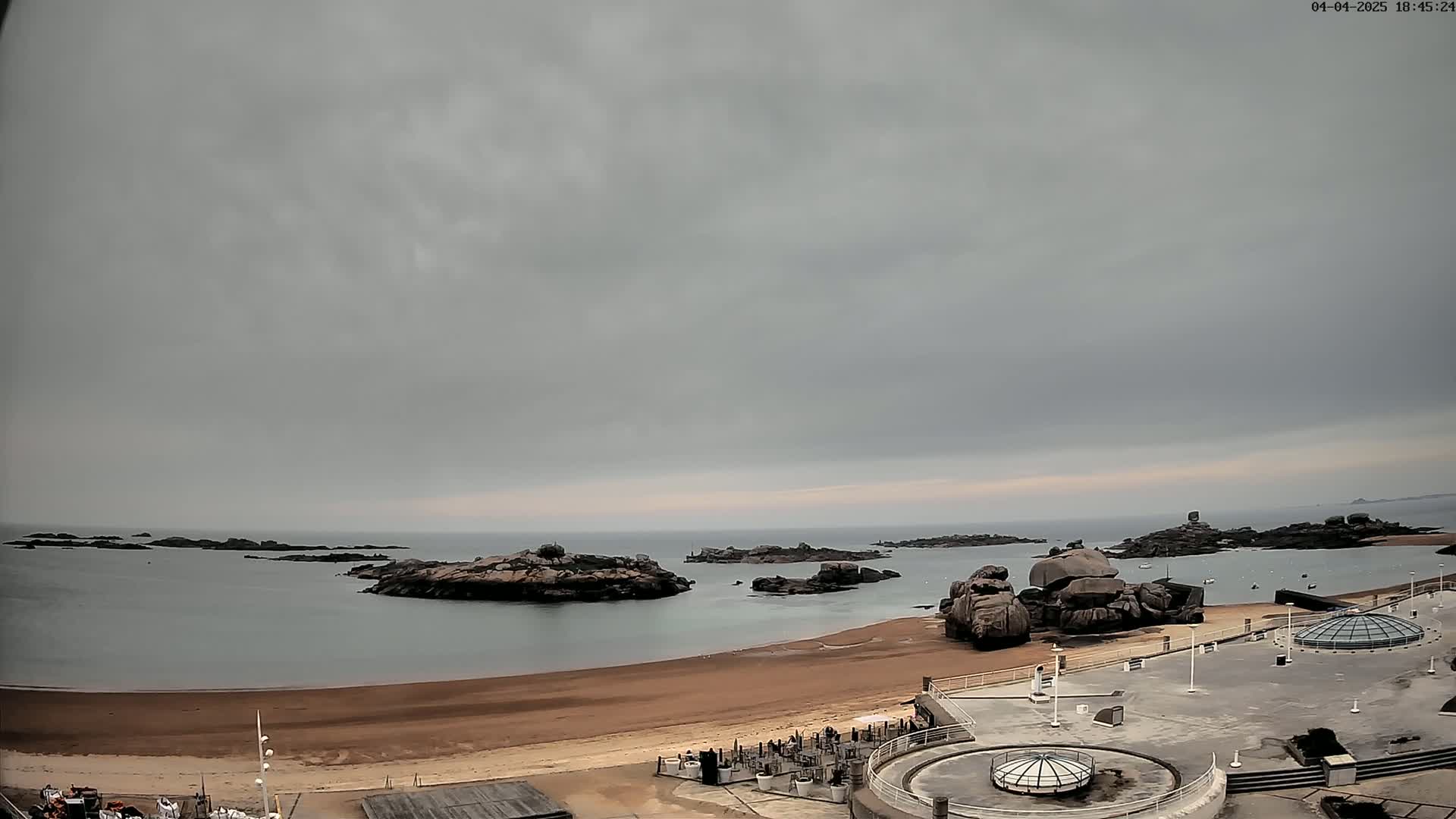 A sandy beach and calm bay are overlooked by a paved area with several dome-shaped structures under an overcast sky.