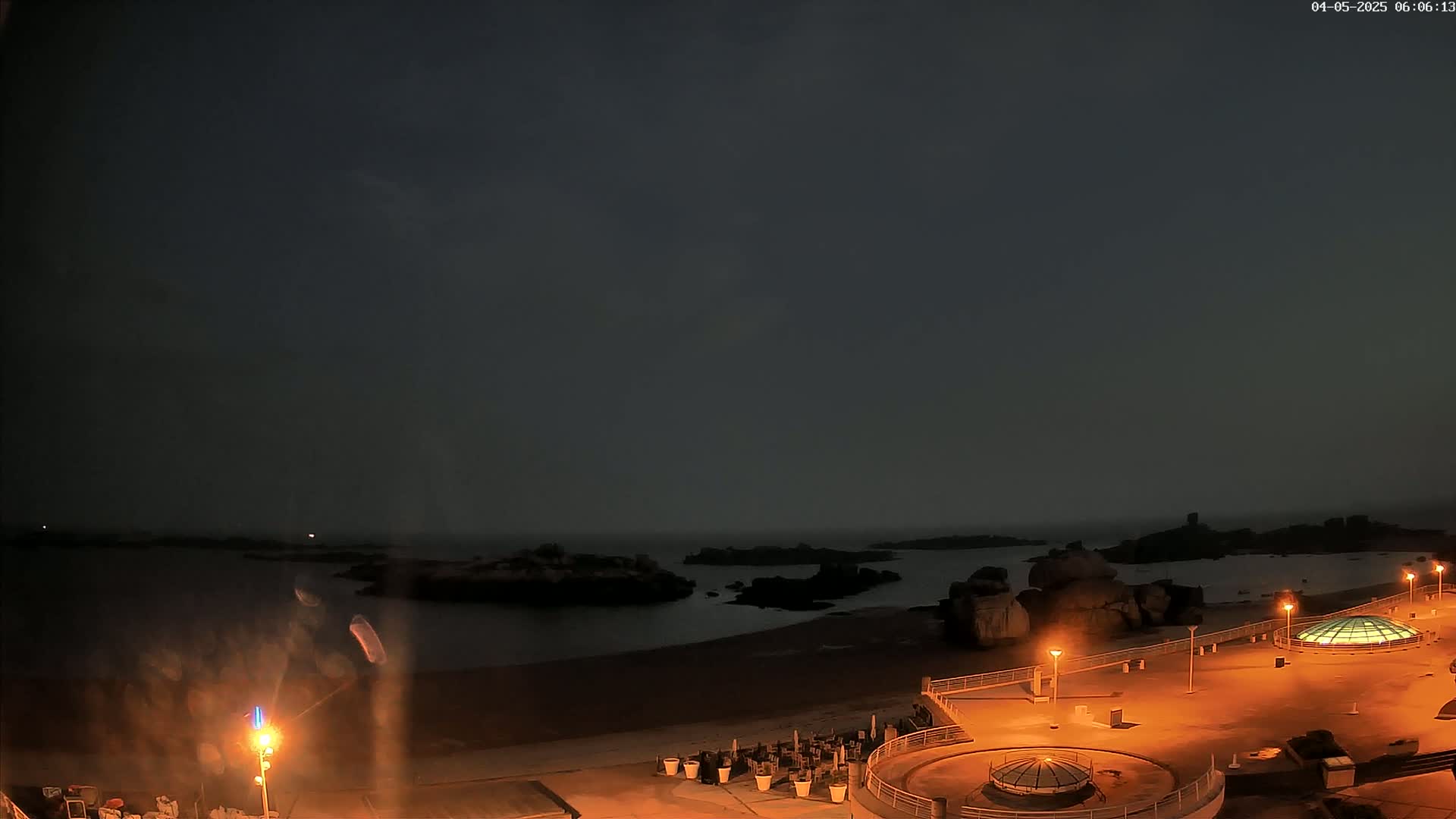A nighttime view of a dark, sandy beach with rocky outcrops in the water, illuminated by artificial lights, under a hazy, dark sky.