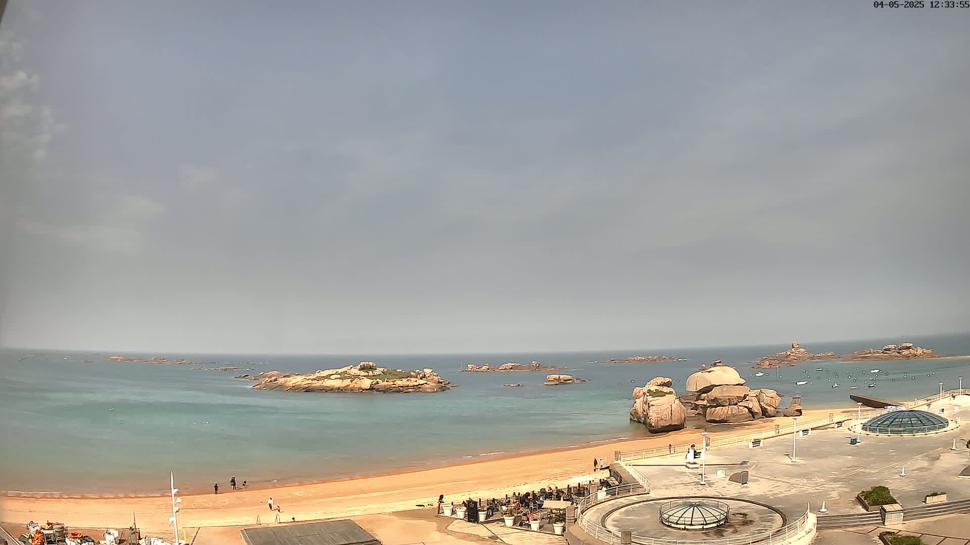 A sandy beach with several people on it is next to a calm, light-blue ocean with rocky islands and a paved area with structures.