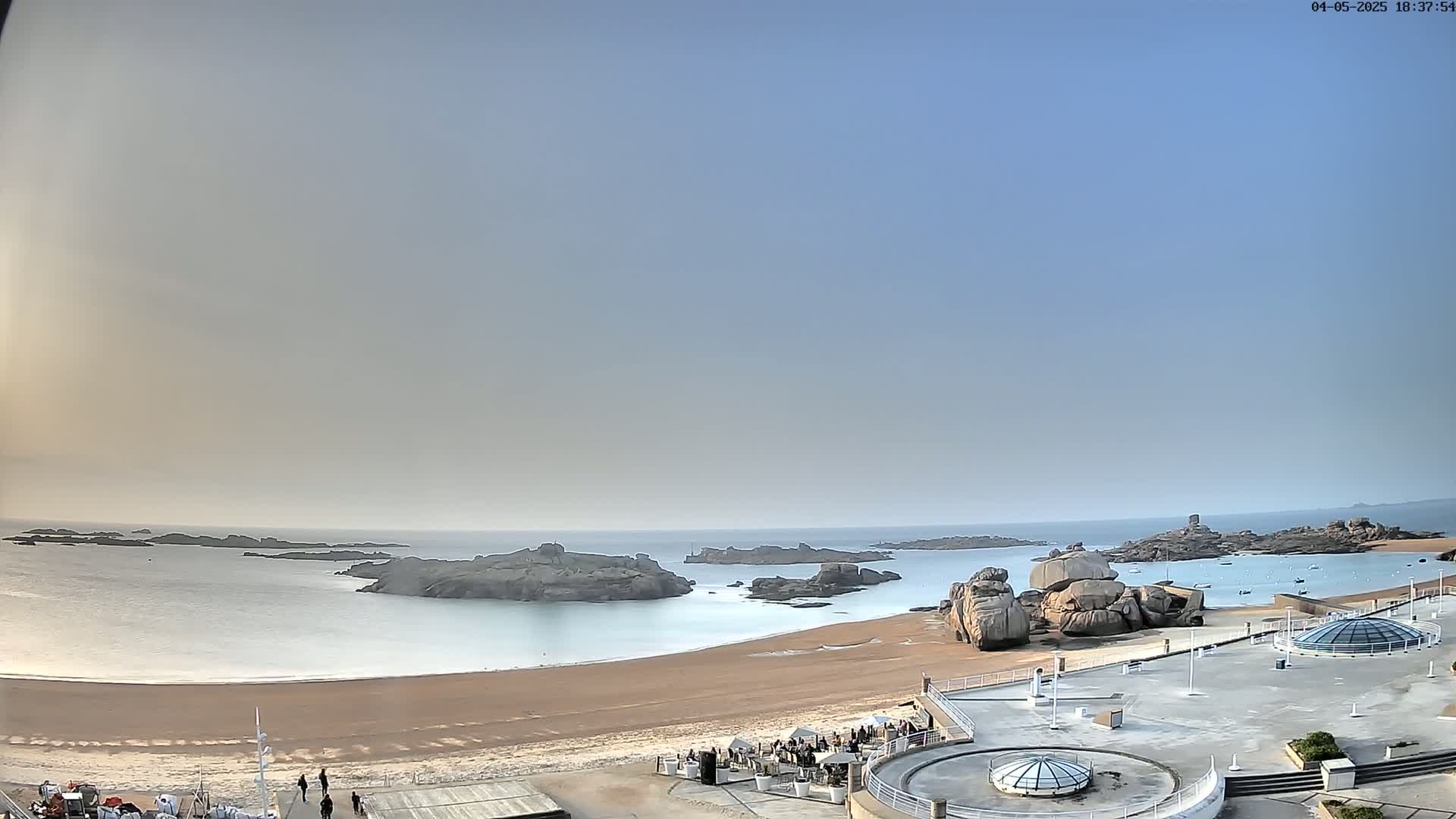 A sandy beach with calm, light-blue water curves around rocky outcroppings under a clear, light-blue sky.