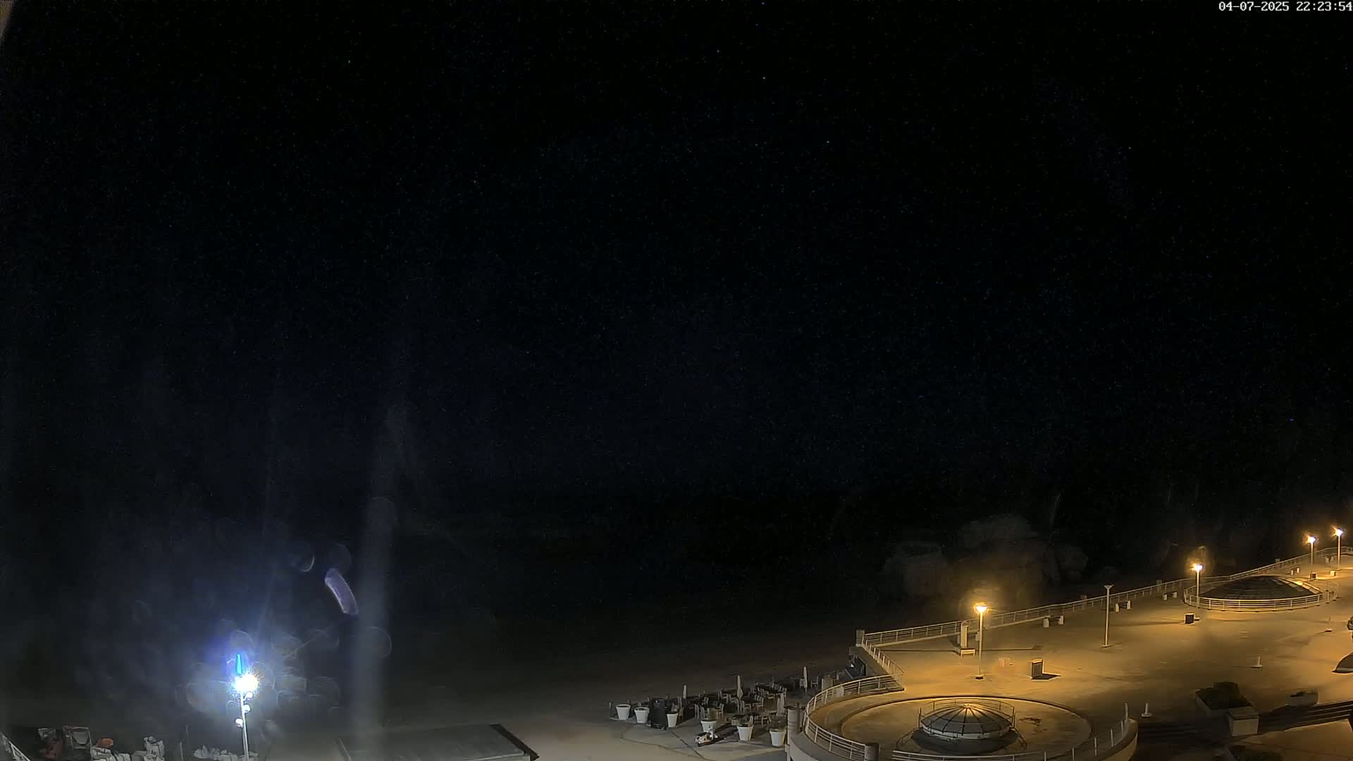 A nighttime, outdoor view of a mostly empty plaza area next to a body of water under a clear, starry sky.