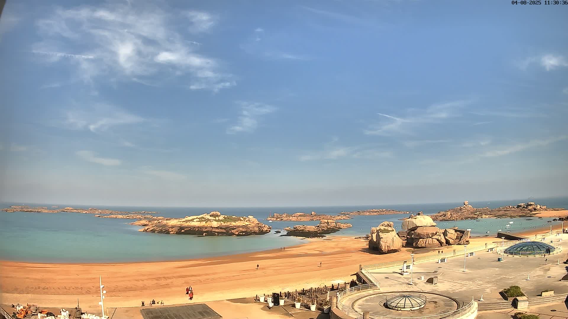 A sandy beach with rocky outcroppings in the ocean under a mostly clear, bright blue sky.