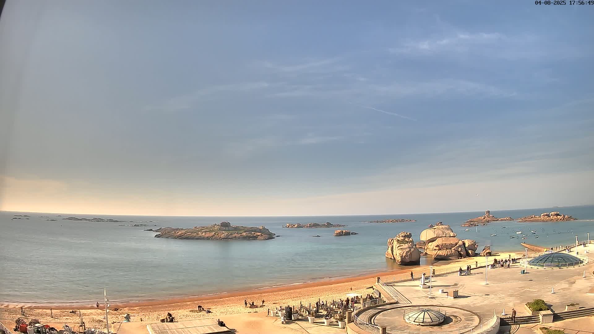 A sandy beach with a few people on it is next to calm blue water with rocky islands visible in the distance under a mostly clear, light-blue sky.