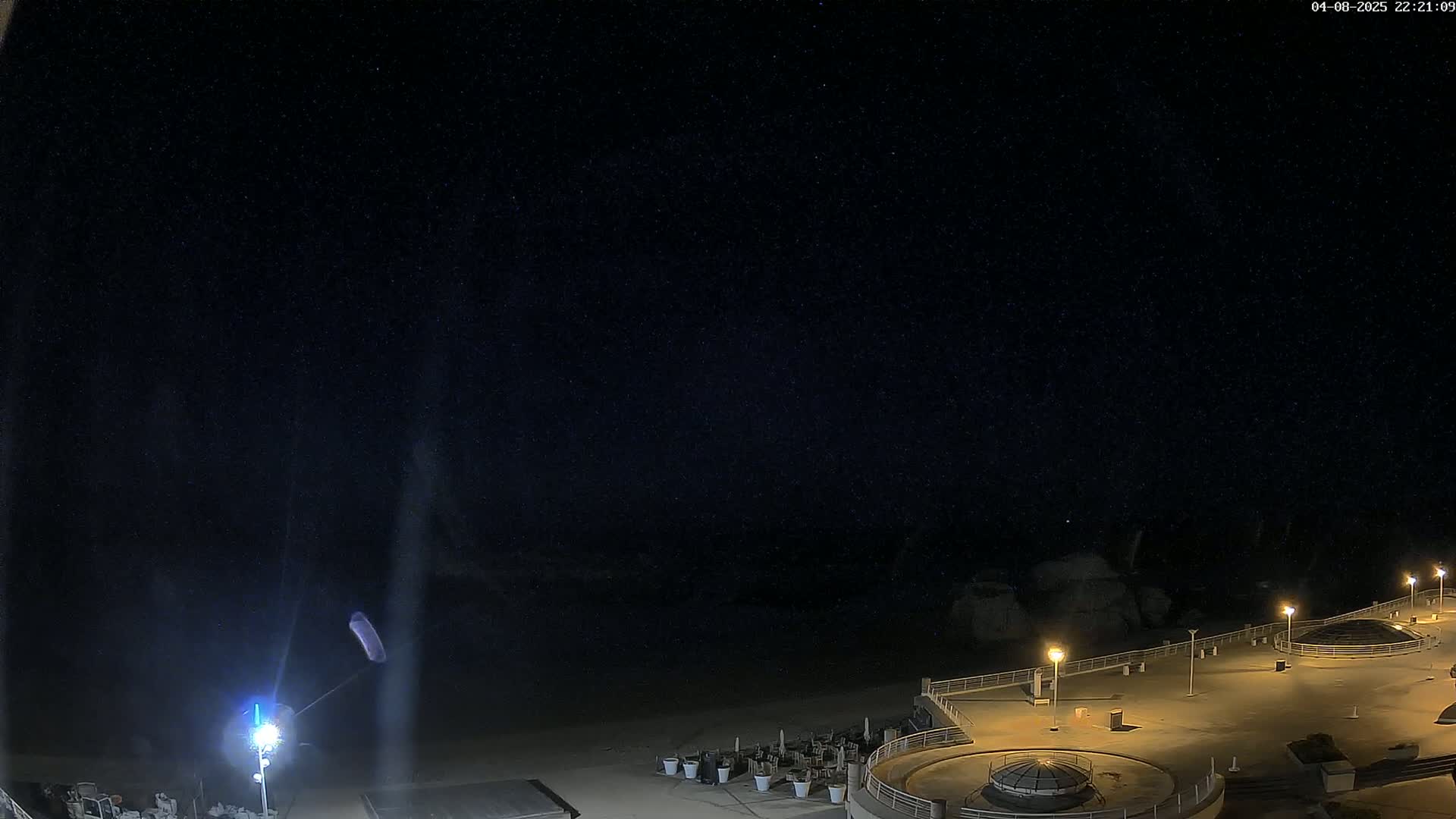 A nighttime, starlit view of a mostly empty beachfront promenade with several lights illuminating the area.