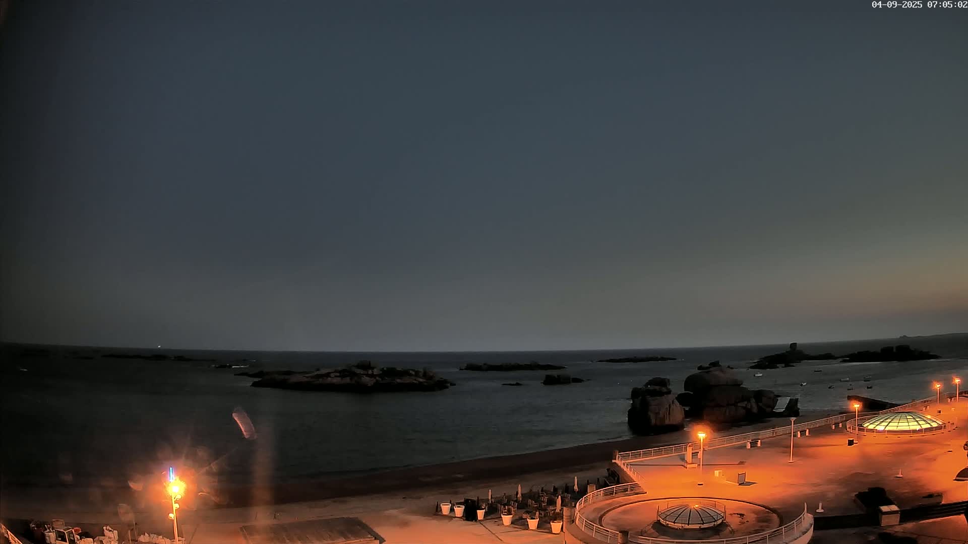 A dark, mostly clear night shows a rocky coastline with a beach, illuminated by artificial lights from a nearby structure.