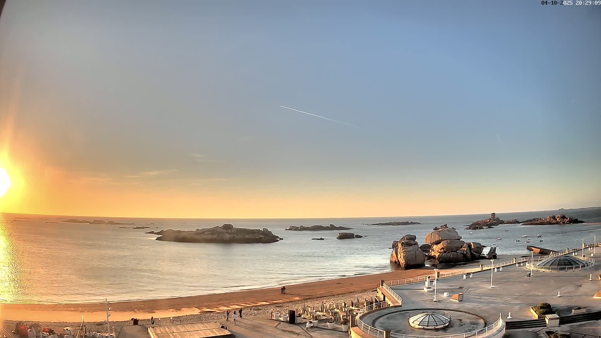 A sunset over a calm ocean with rocky islands and a sandy beach, viewed from a coastal plaza.